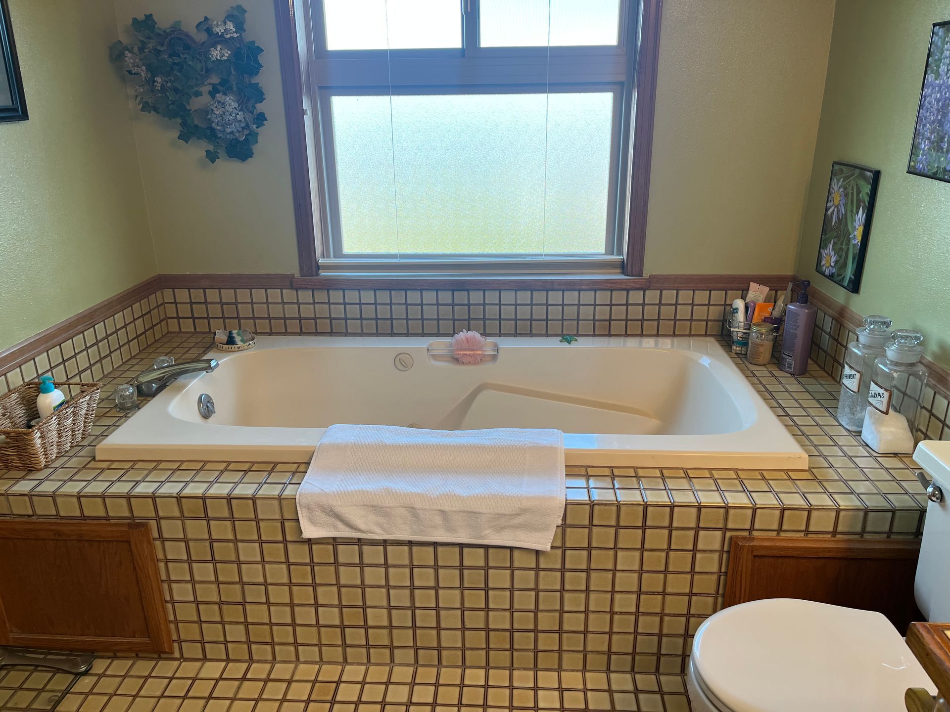 Tiled bathroom with a corner soaking tub beneath a window and a towel draped over the edge