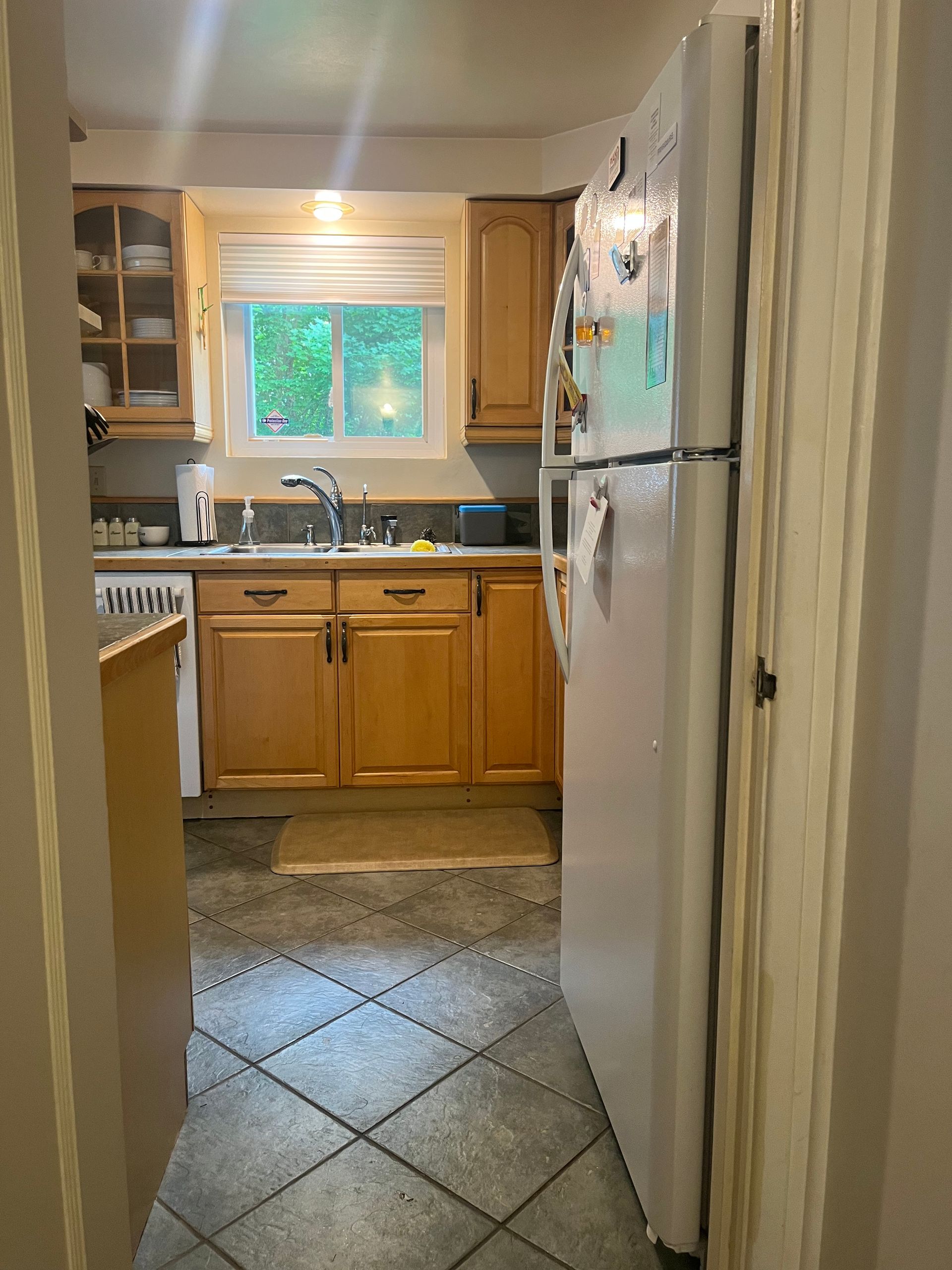 View into a small kitchen with wooden cabinets, white refrigerator, and a window above the sink.