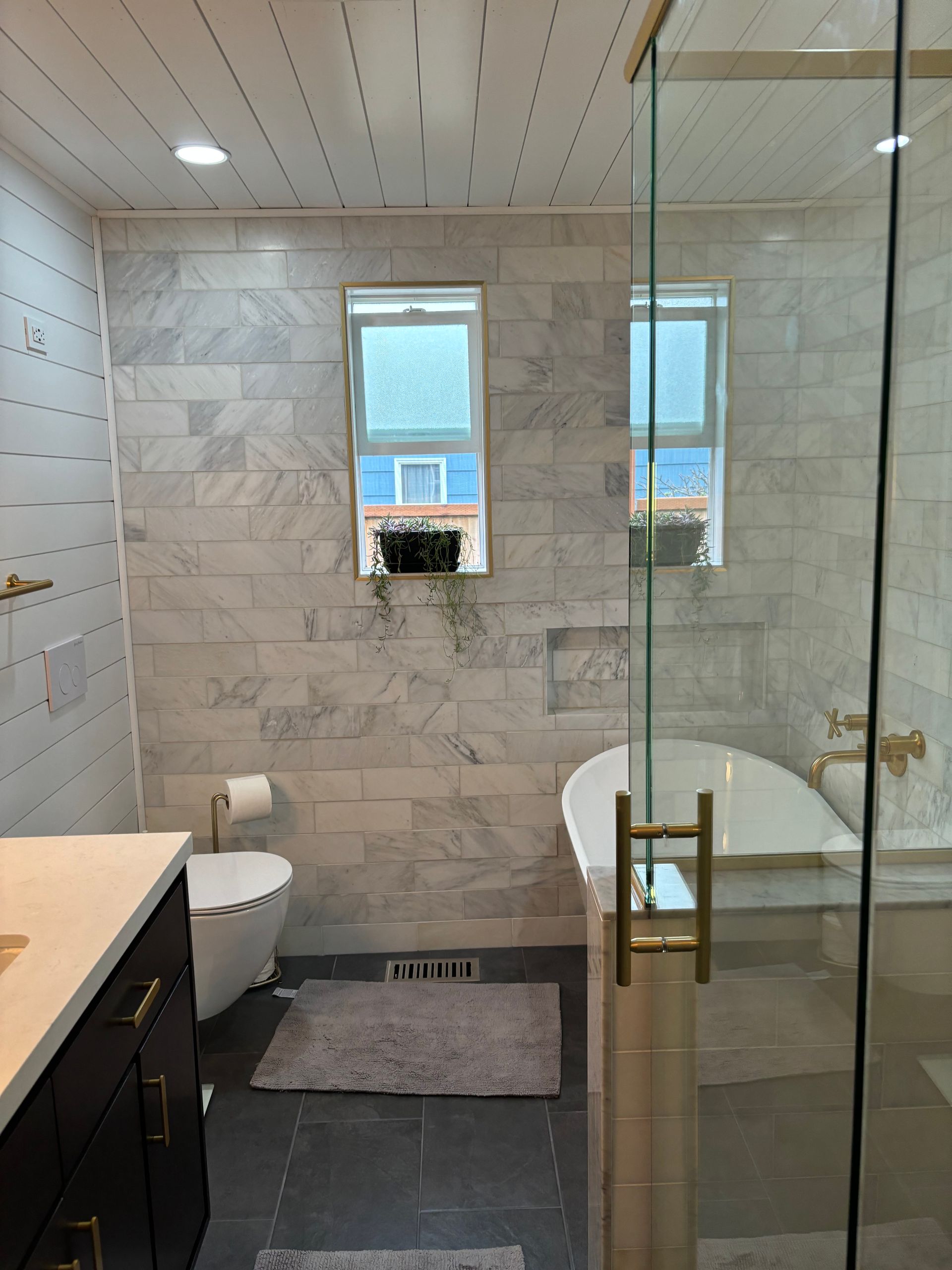 Modern bathroom with tiled walls, glass shower, toilet, and small window overlooking water.