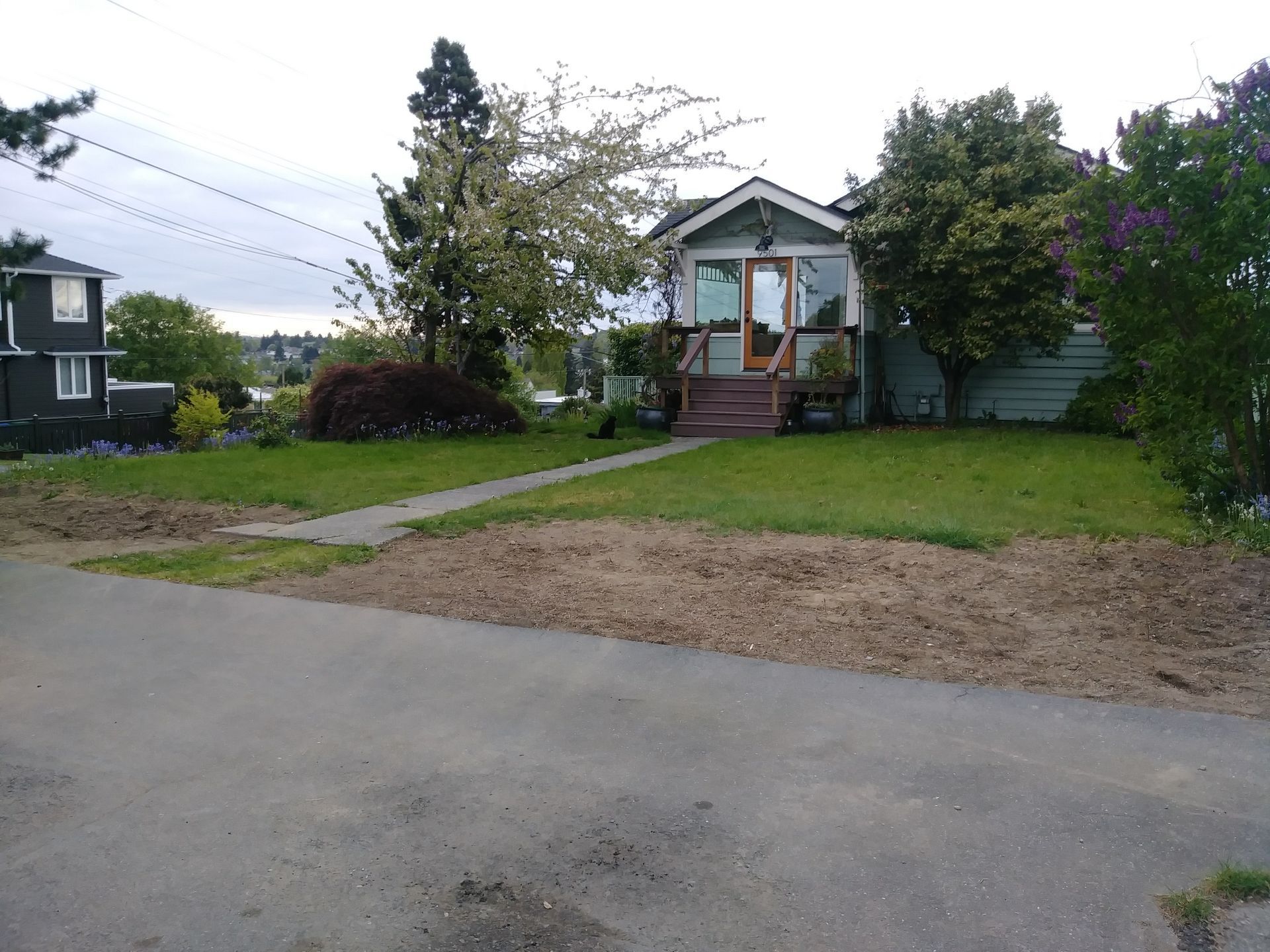 Small house with a front porch and stairs, seen across a gravel road with a grassy yard and trees.