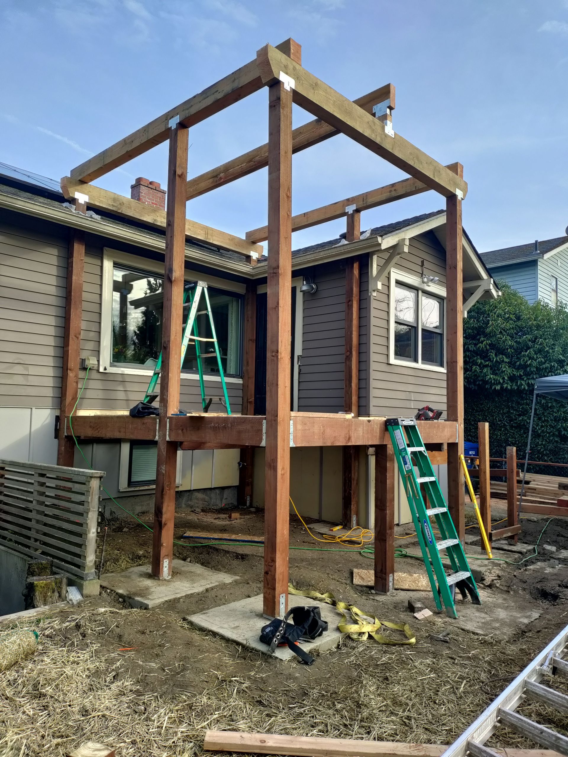 Partially built wooden deck frame beside a house, with green ladders and construction tools in a backyard.