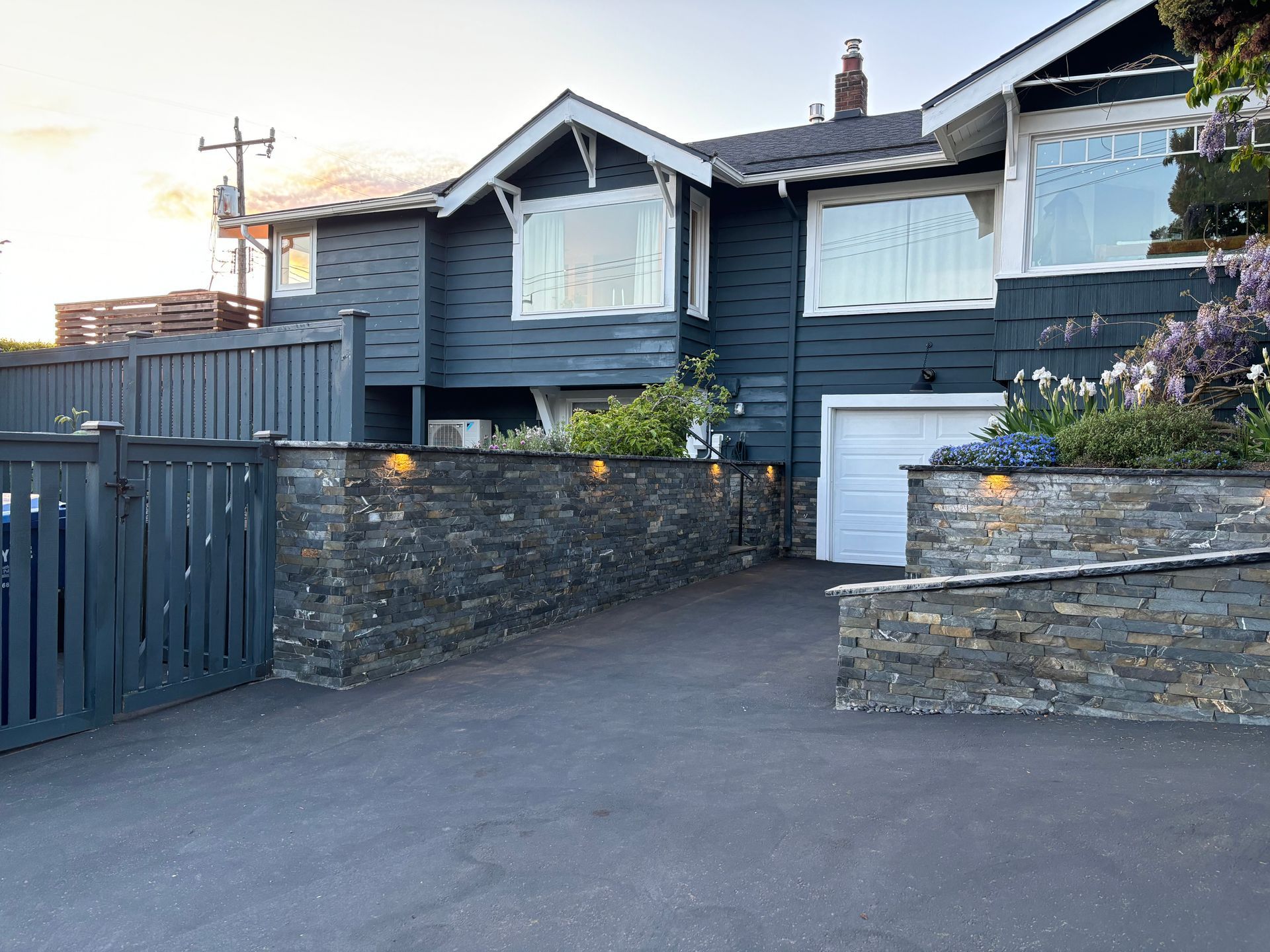 Dark gray house with white-trimmed windows, stone walls, and a driveway at sunset.