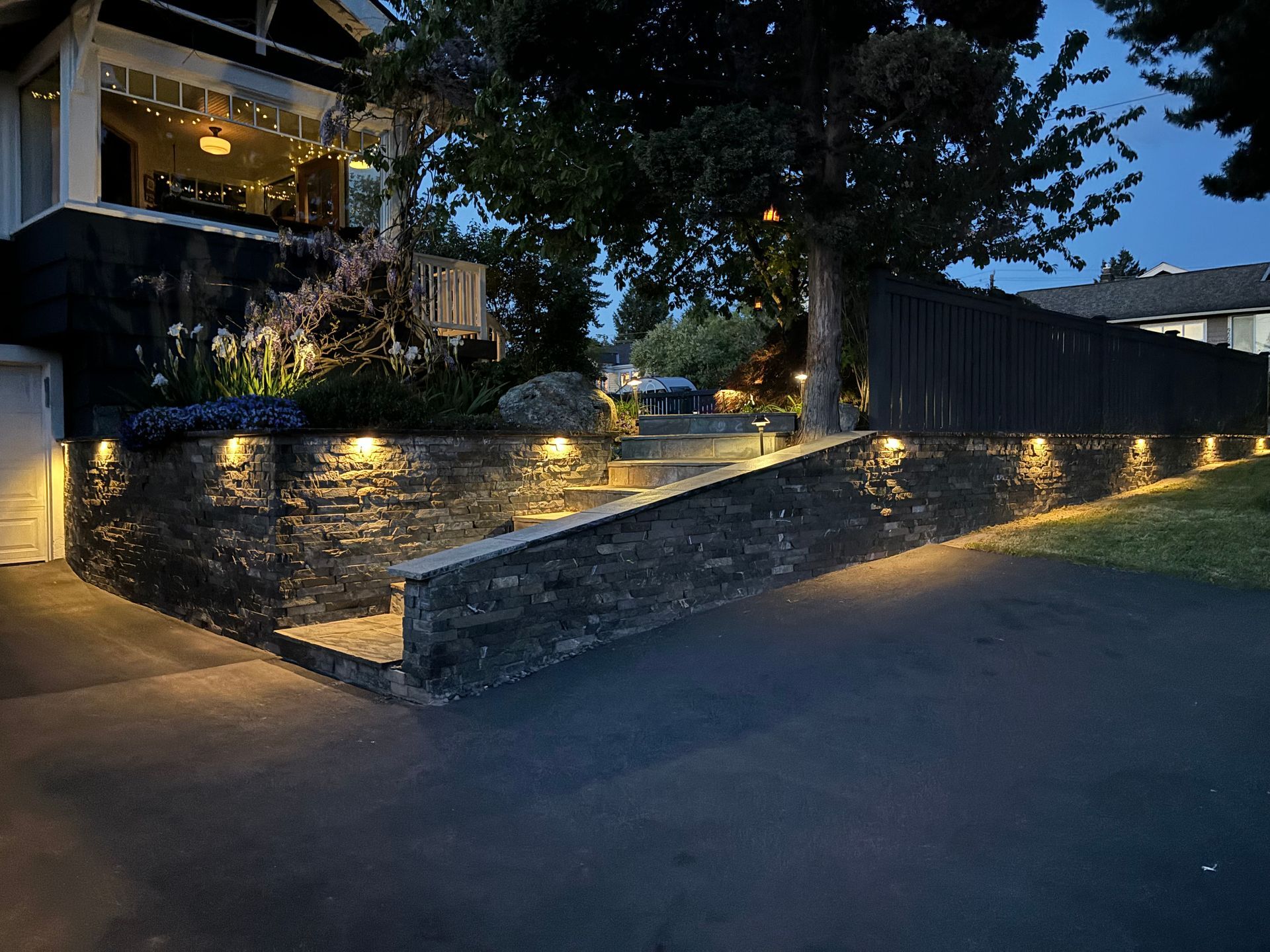 Stone retaining wall and steps lit by warm lights beside a house at dusk