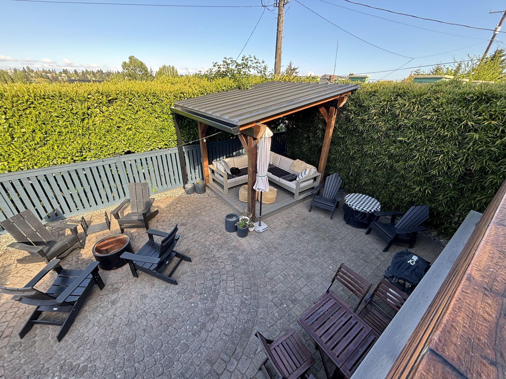 Patio with picnic tables, chairs, and a small covered shelter beside tall hedges and a gravel ground