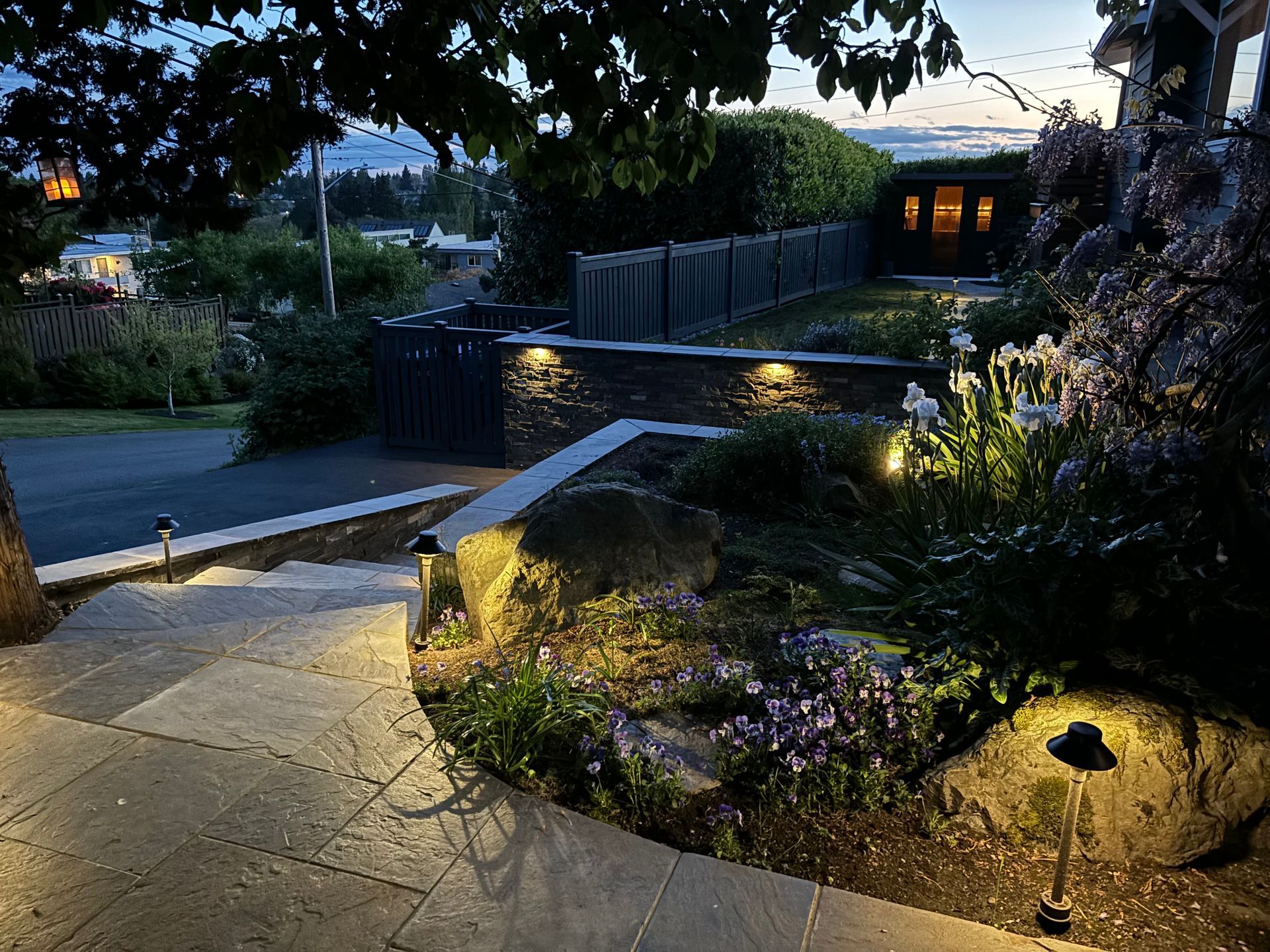 Lit garden walkway and stone steps beside a house at dusk, with shrubs and warm lights.