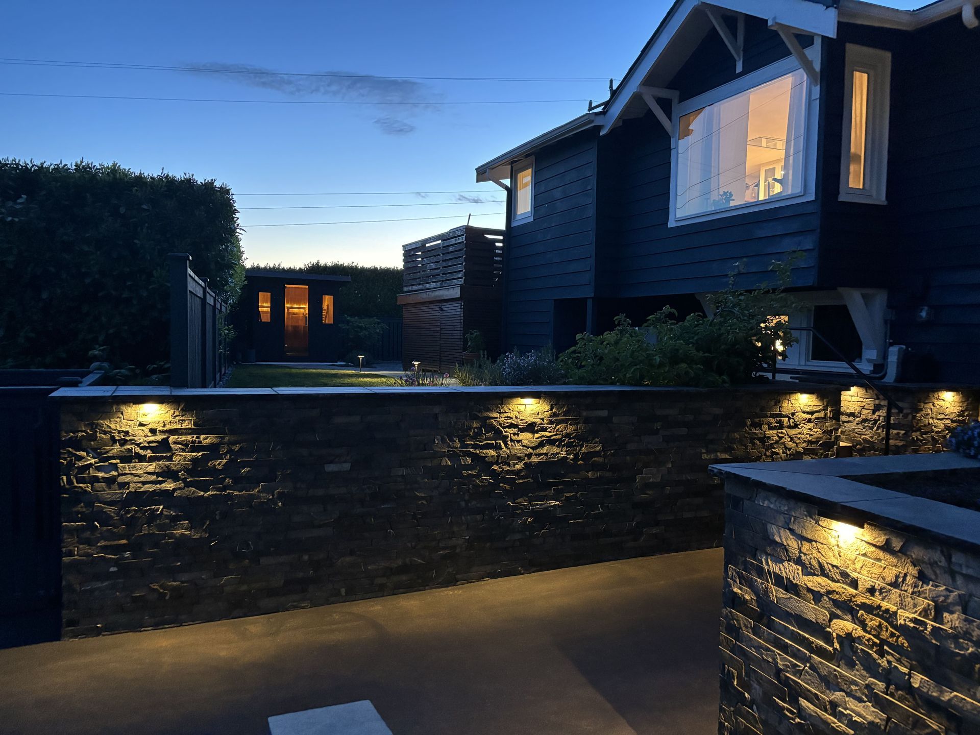 Modern house exterior at dusk with lit stone wall and blue evening sky