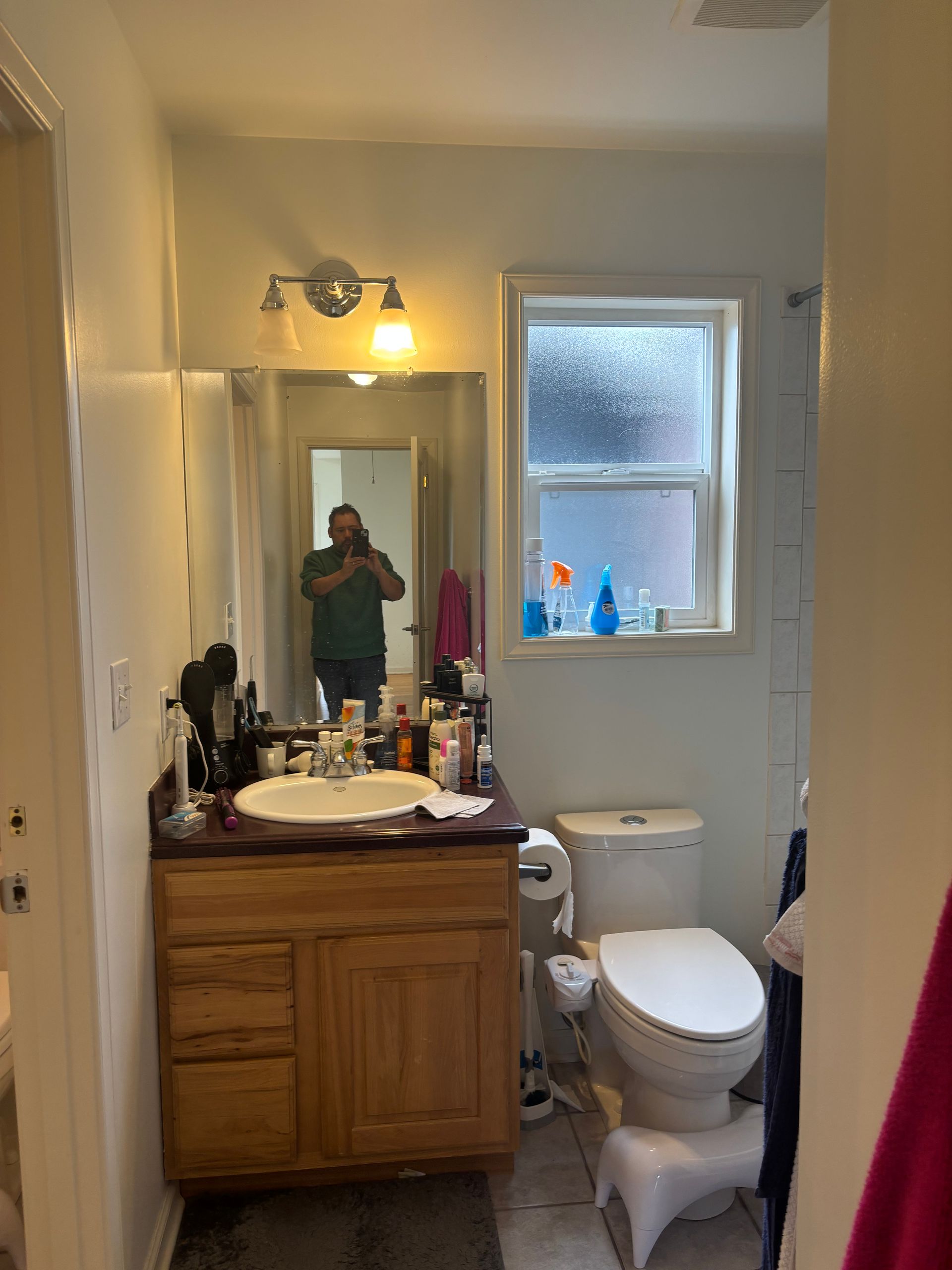 Small bathroom with wood vanity, mirror, toilet, and a frosted window; person visible in mirror.