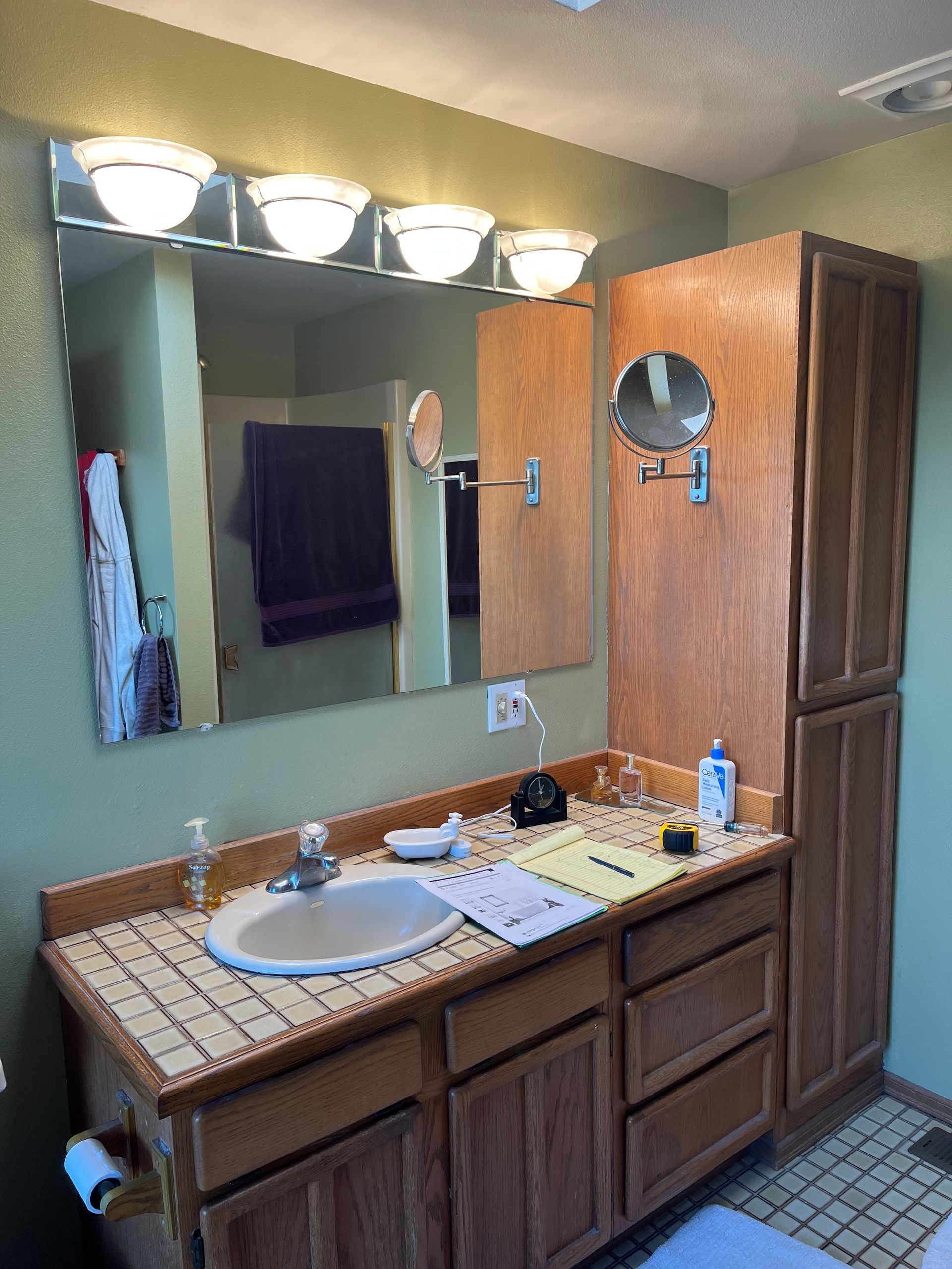 Bathroom vanity with sink, mirror, lights, and brown cabinets; tiled floor and purple towel hanging nearby