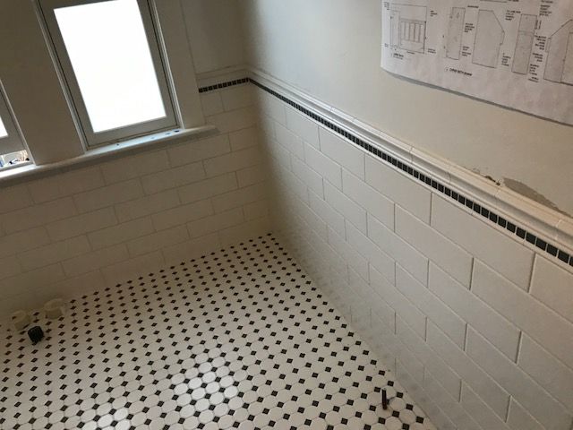 White-tiled bathroom with black-and-white dotted floor and a window on the left.
