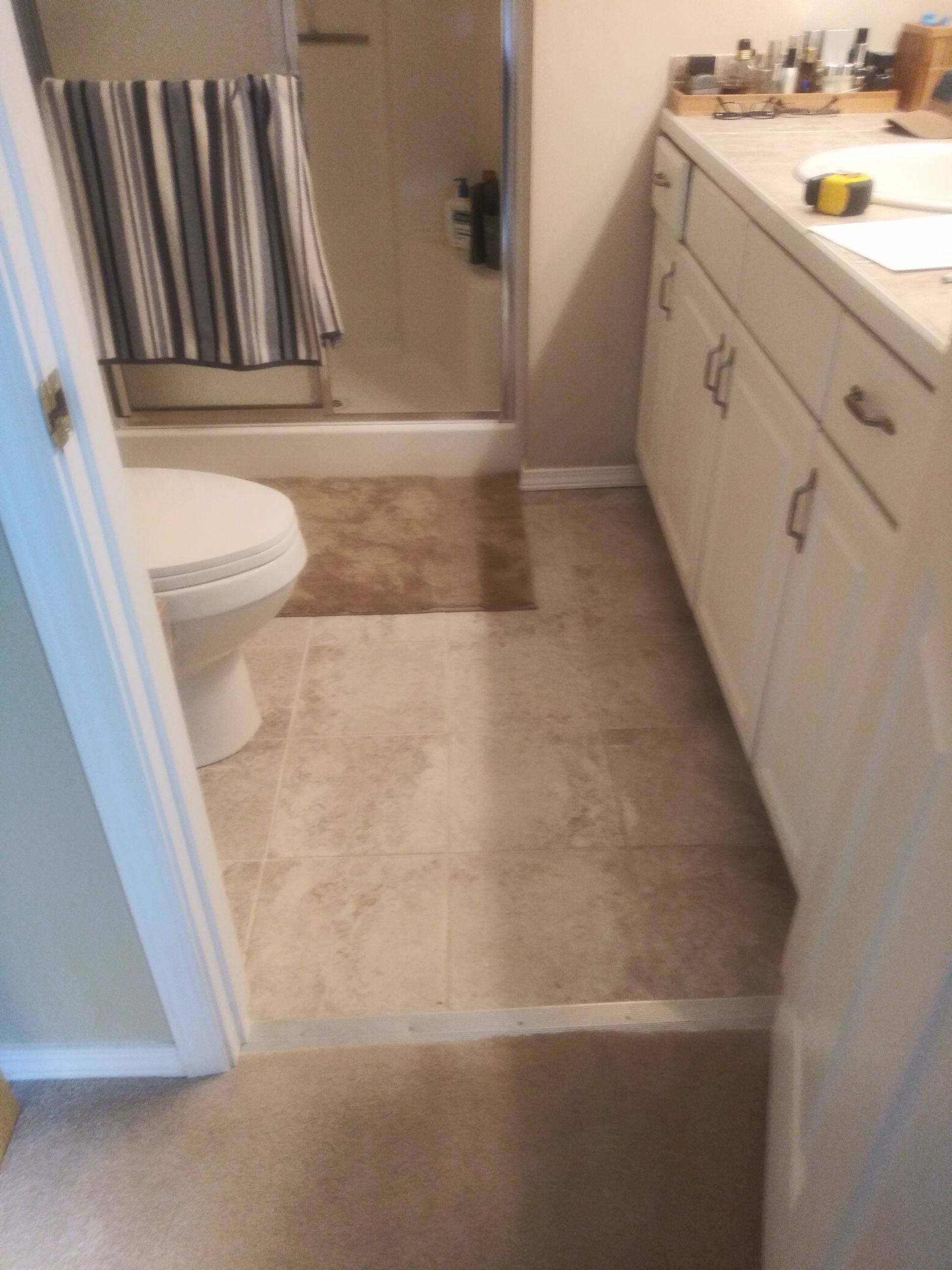 Bathroom with tan tile floor, white vanity, toilet, and shower curtain near a tiled shower stall.