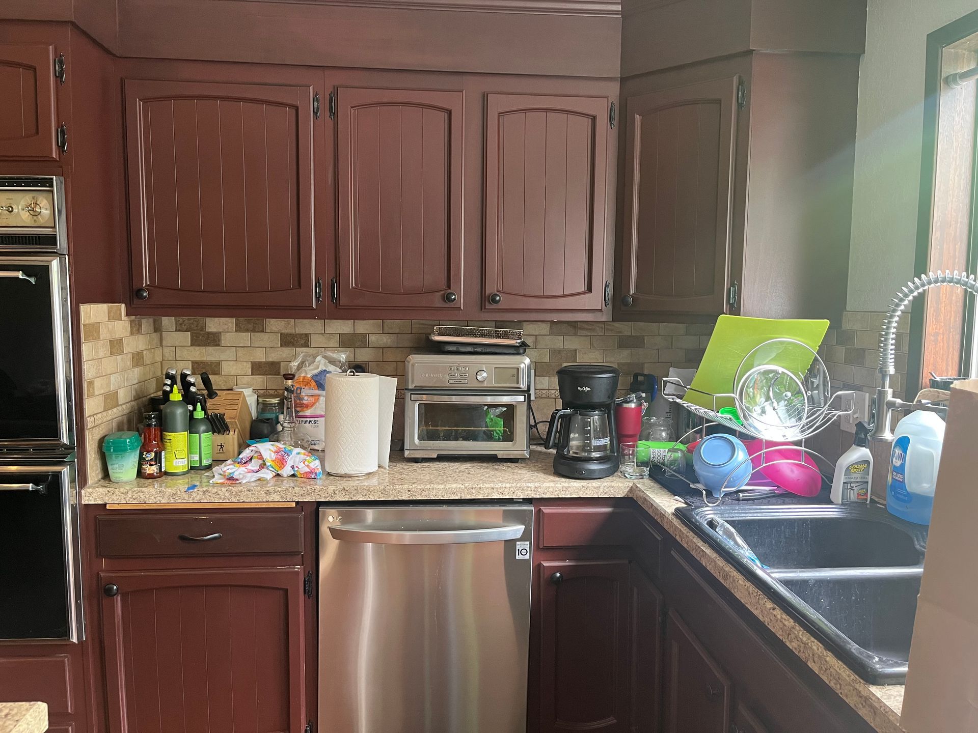 Compact kitchen with dark wood cabinets, stainless dishwasher, sink, and countertop items by a window