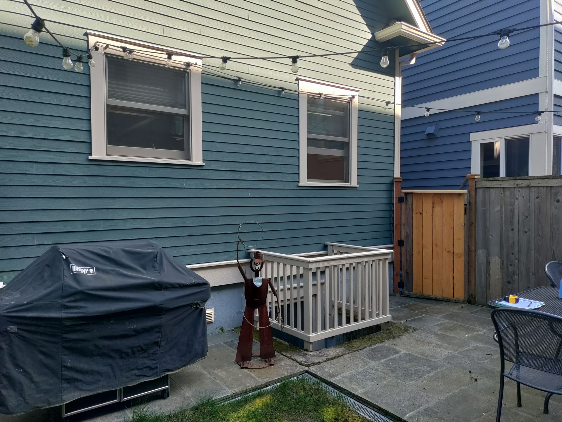 Backyard patio beside a blue house with a covered grill, string lights, and a small white railing.