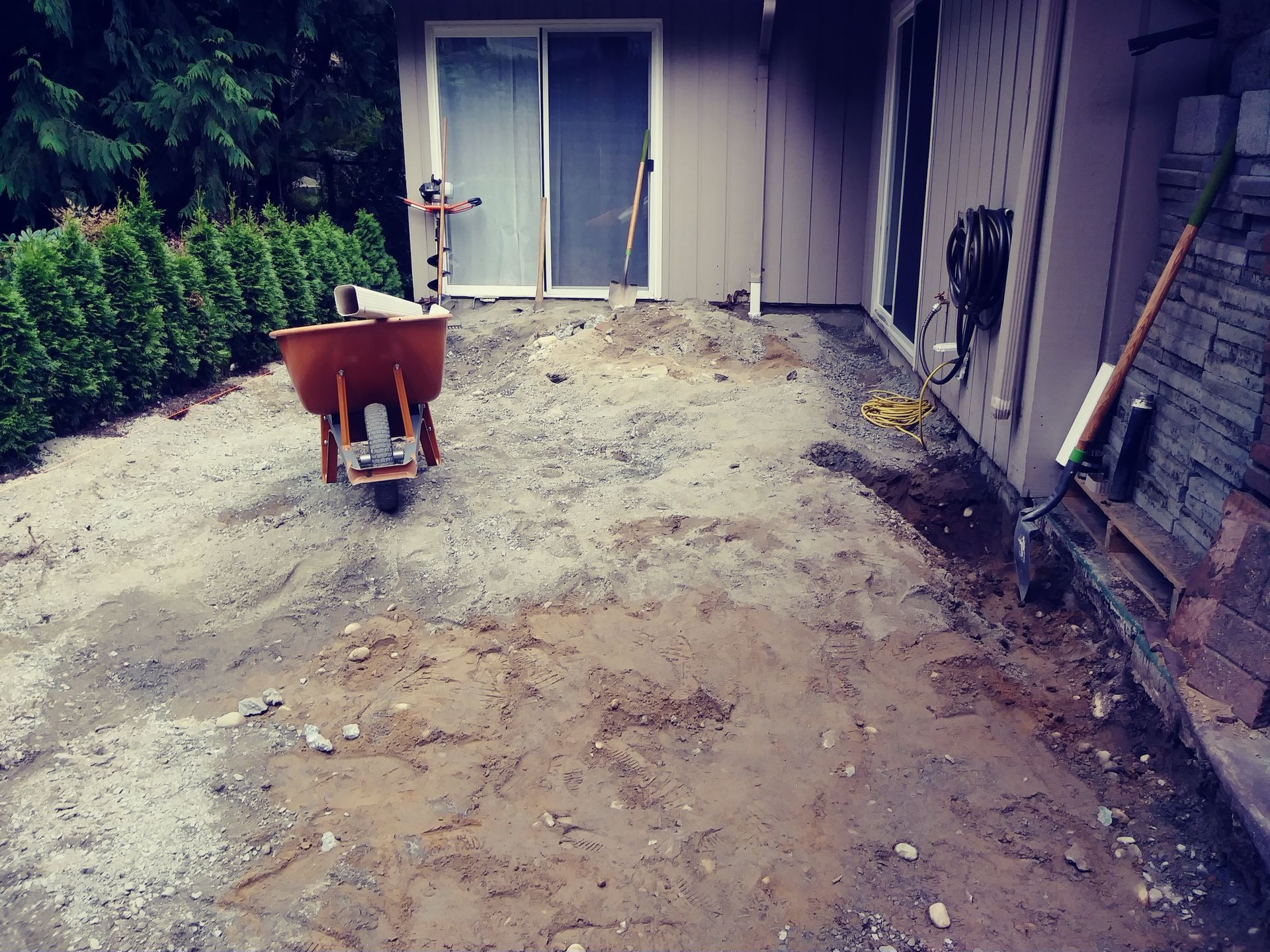 A muddy backyard with a wheelbarrow near a sliding glass door and house siding.