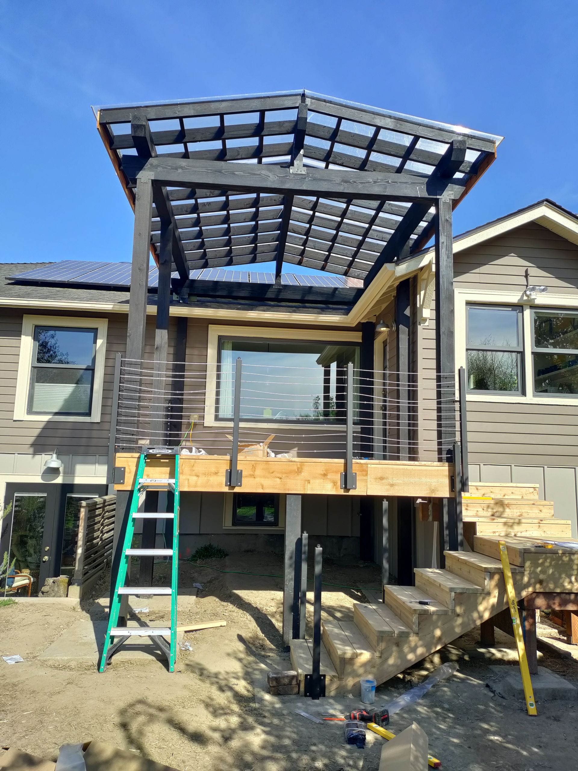 House under construction with a raised deck, pergola frame, and ladders in a backyard.