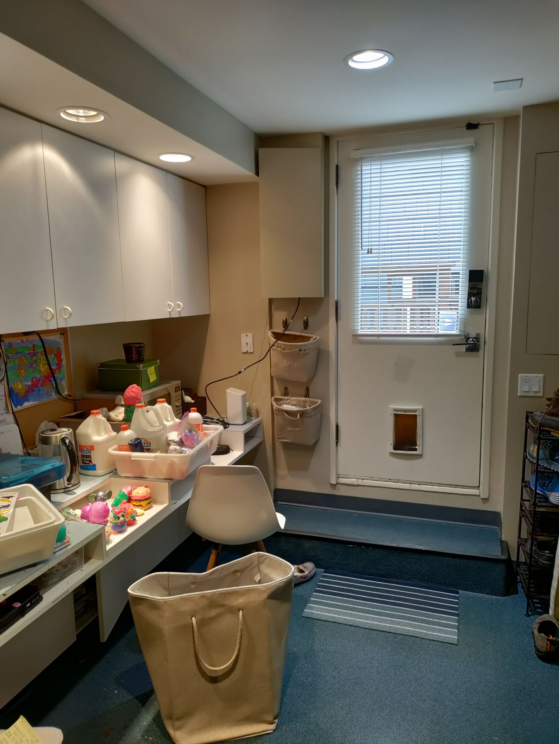 Small craft room with white cabinets, cluttered worktable, chair, and blue carpet near a door with window blinds