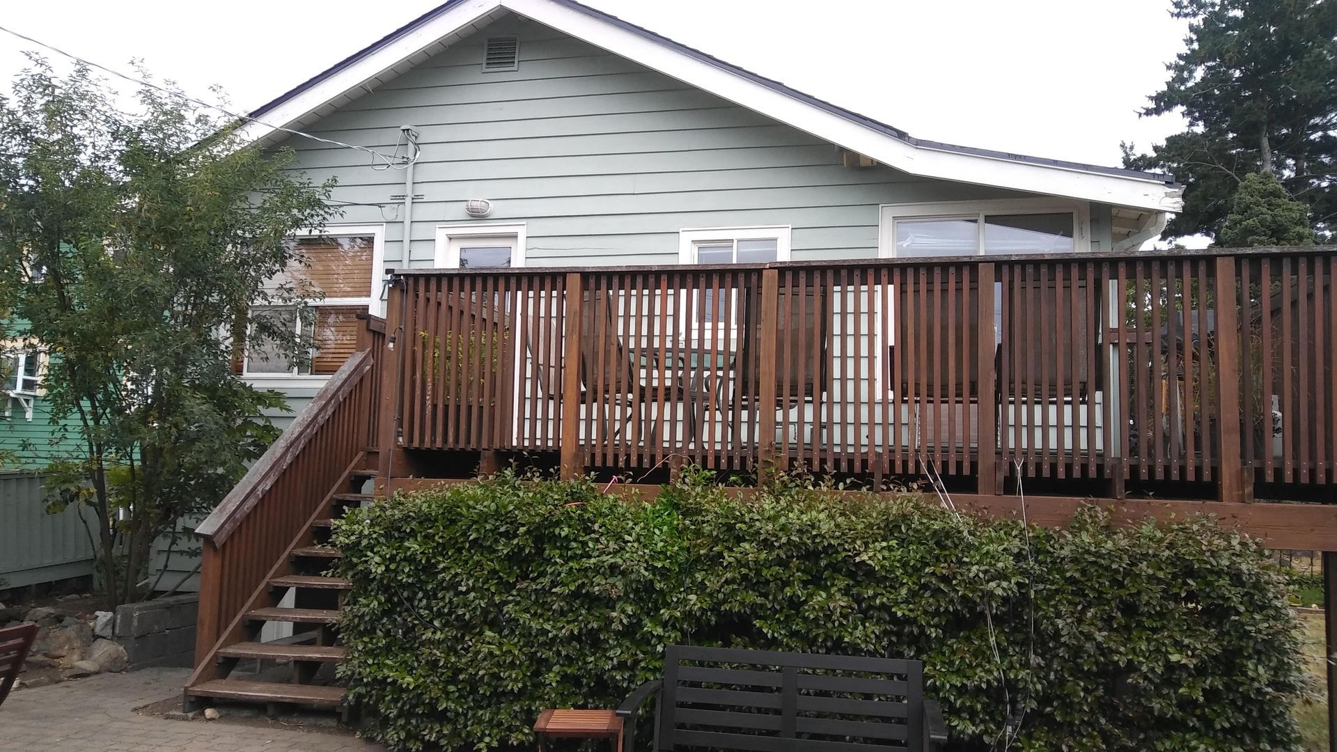 Wooden deck with stairs on the back of a light gray house, partially covered by overgrown bushes.