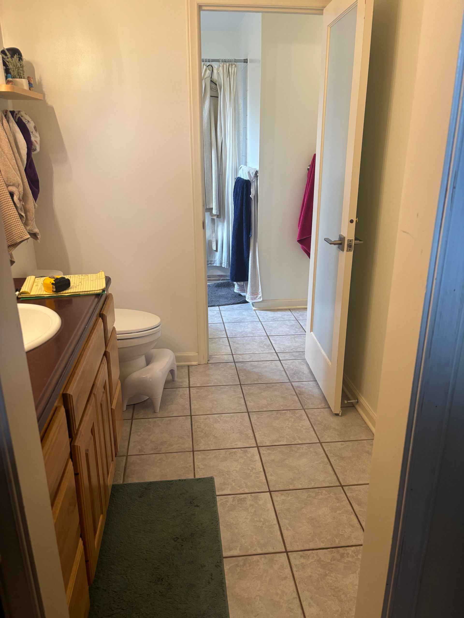 Narrow bathroom with tiled floor, wooden vanity, toilet, and open door leading to a closet or hallway.