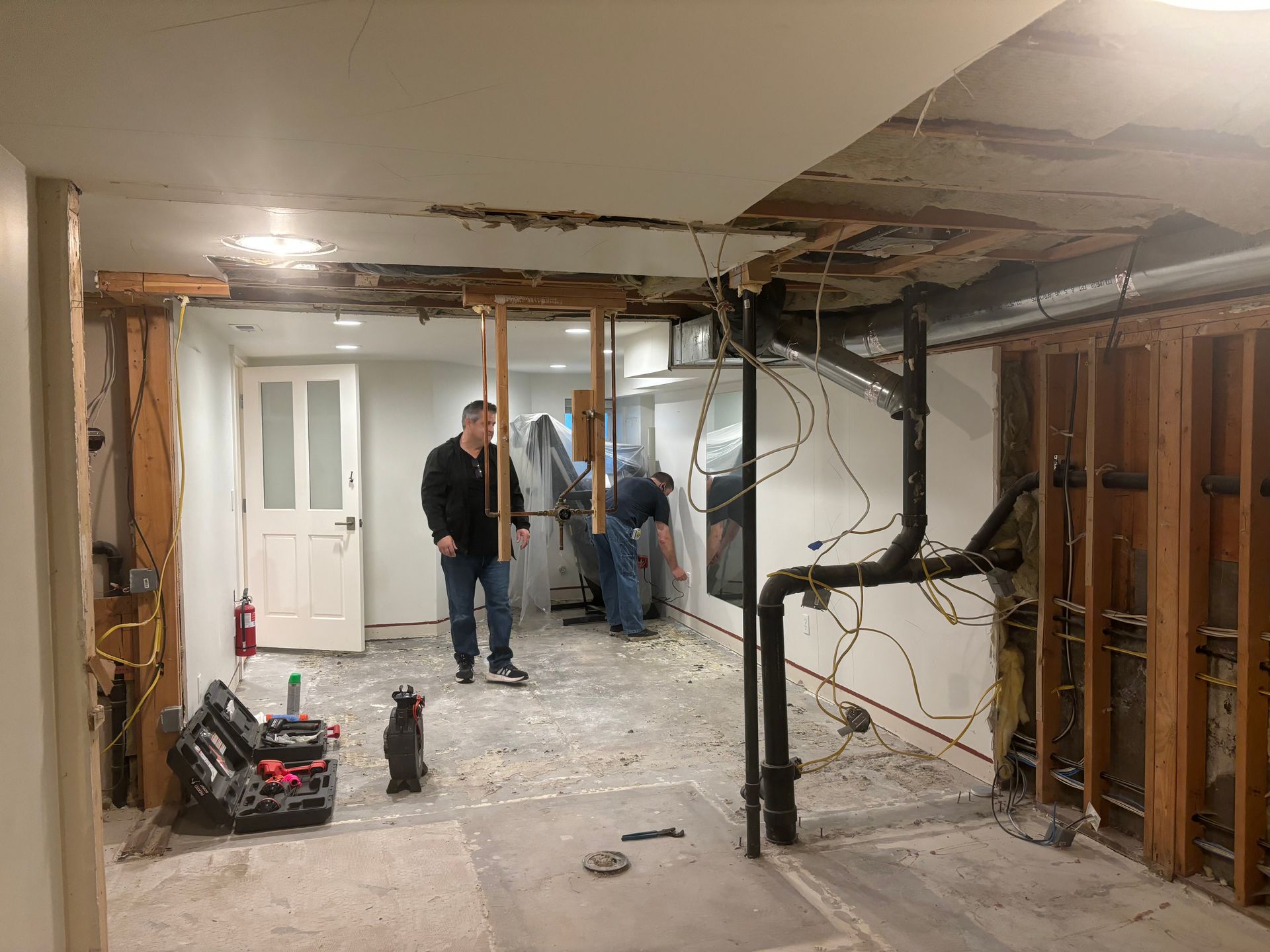 Unfinished basement with exposed studs, debris on floor, and two people working near a doorway.