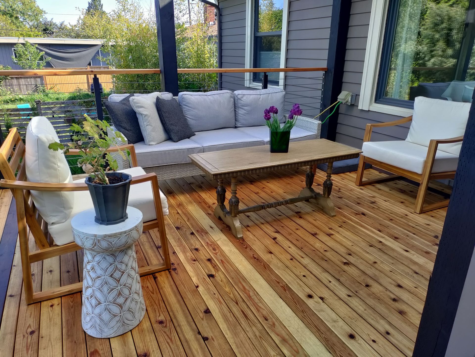 Wooden patio with gray sofa, chairs, coffee table, and potted plants by a glass door
