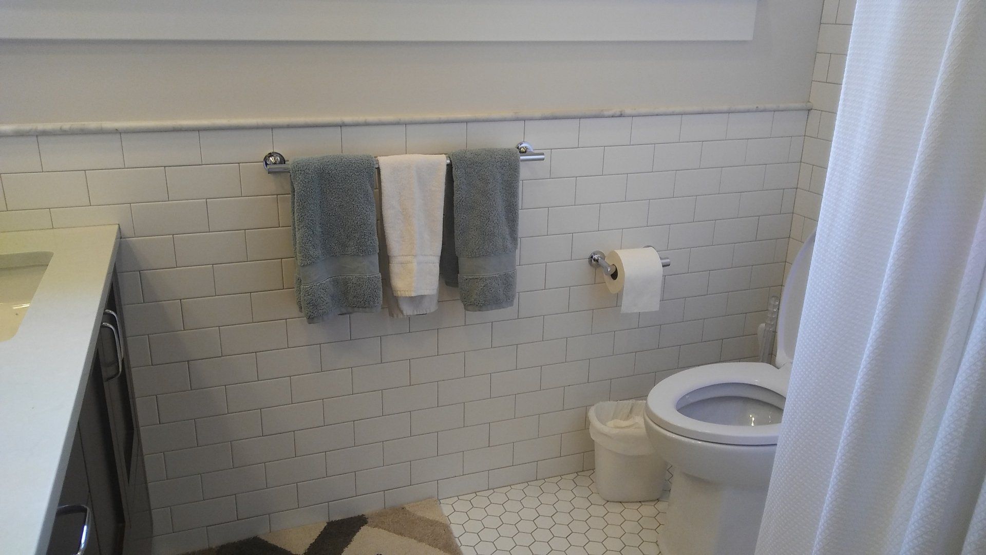 Small bathroom with tiled wall, towels on hooks, toilet paper holder, trash can, and shower curtain.