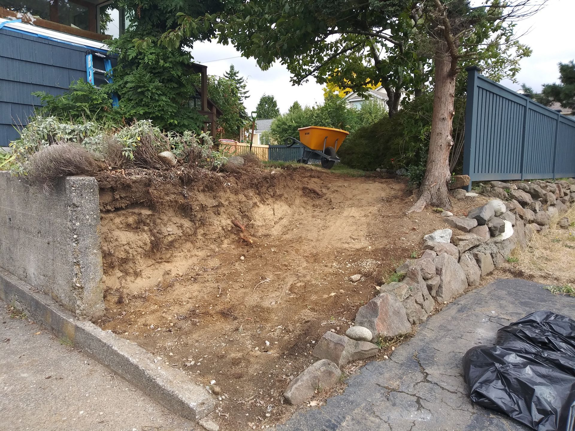 Eroded dirt slope beside a stone retaining wall, with a tree, blue fence, and black tarp in a yard