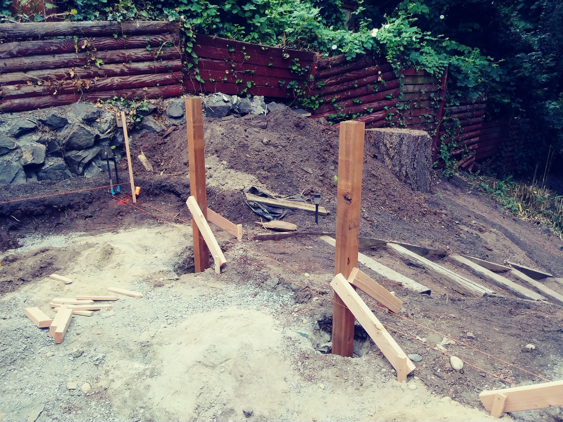 Construction site with two wooden posts in dirt and a gravel area, backed by stacked logs and a stone wall