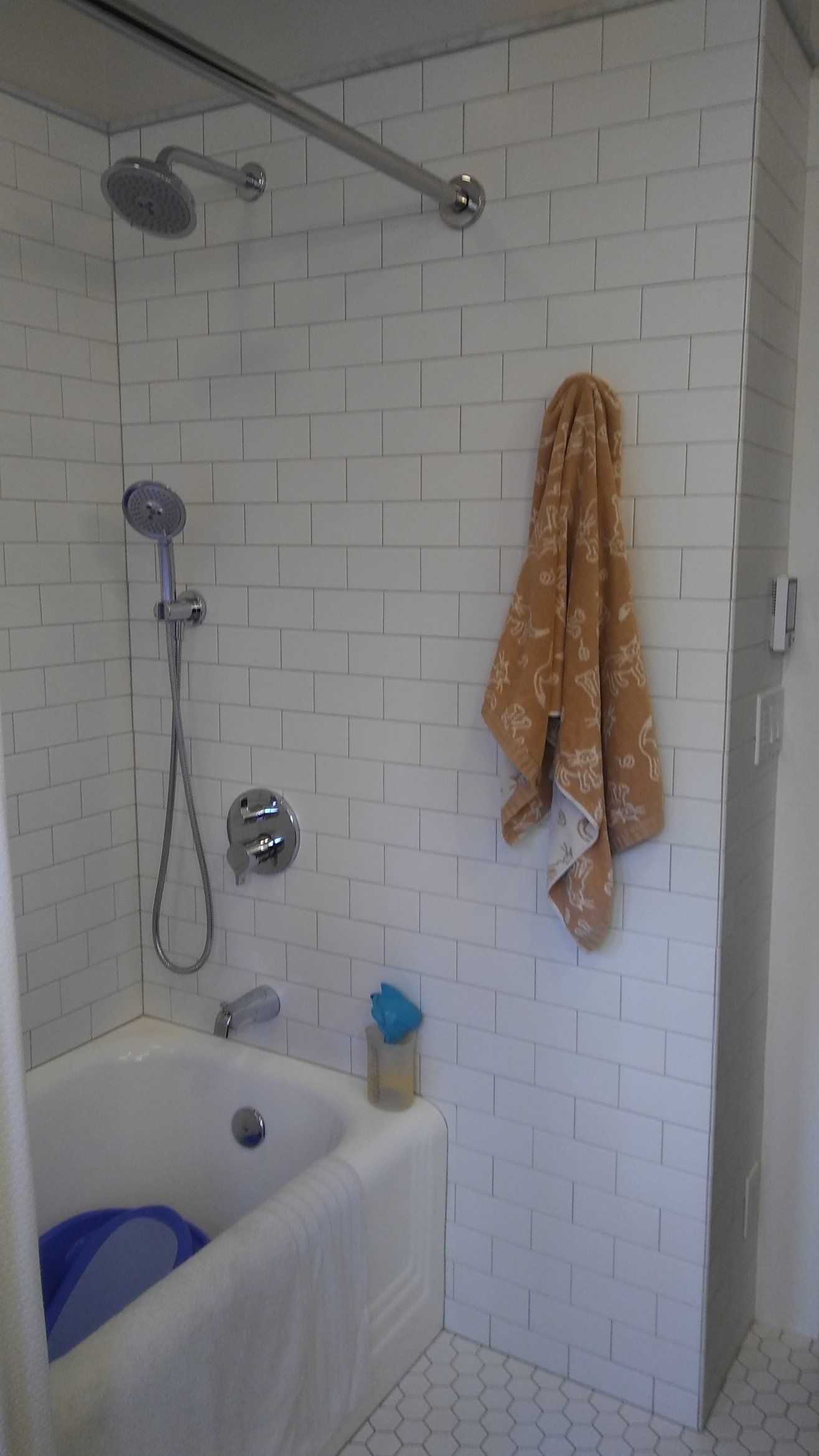 Small white-tiled bathroom shower with a blue tub, handheld showerhead, and orange towel on the wall