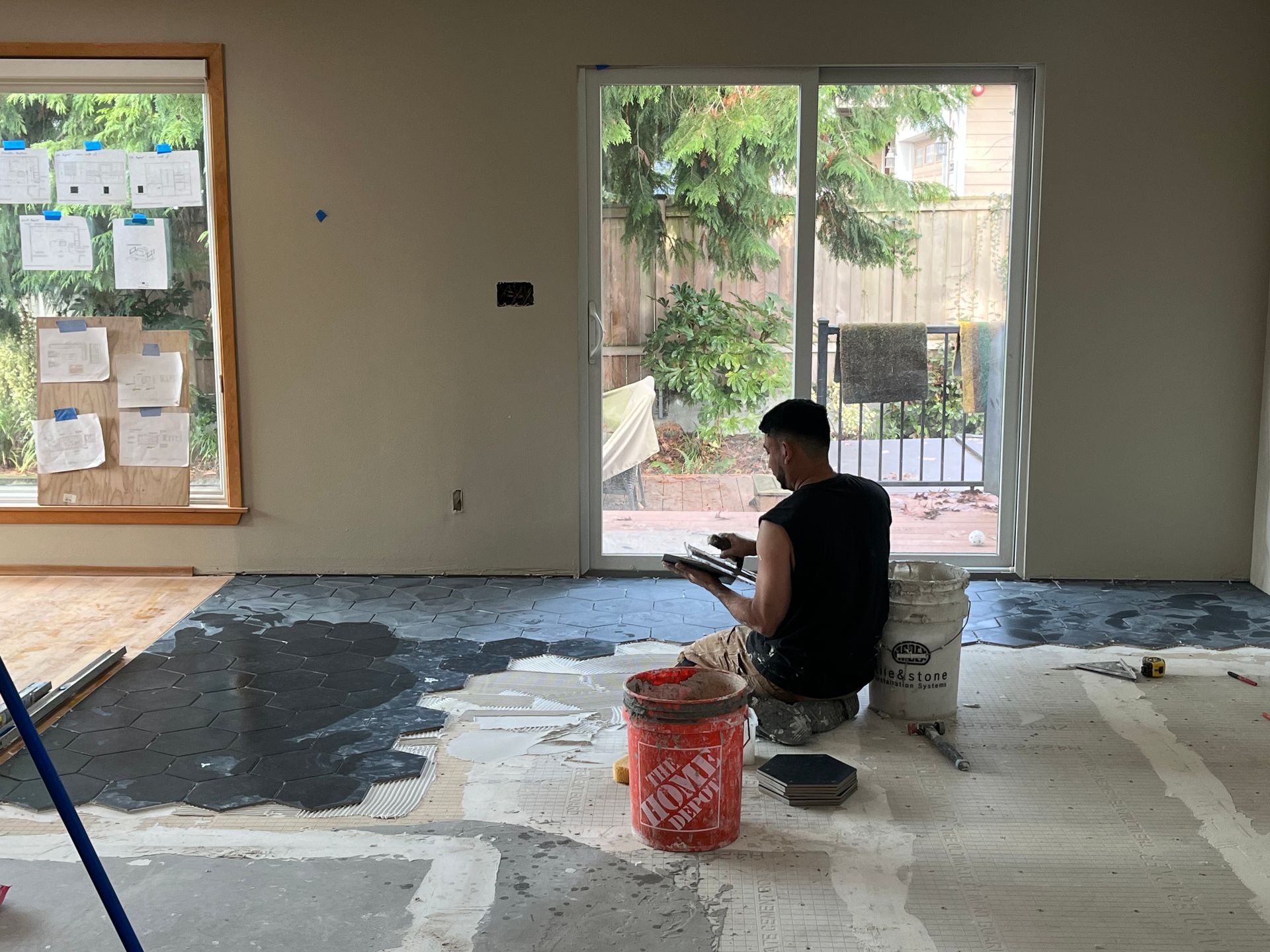 Worker sitting on the floor in an unfinished room, applying or mixing material near open windows.