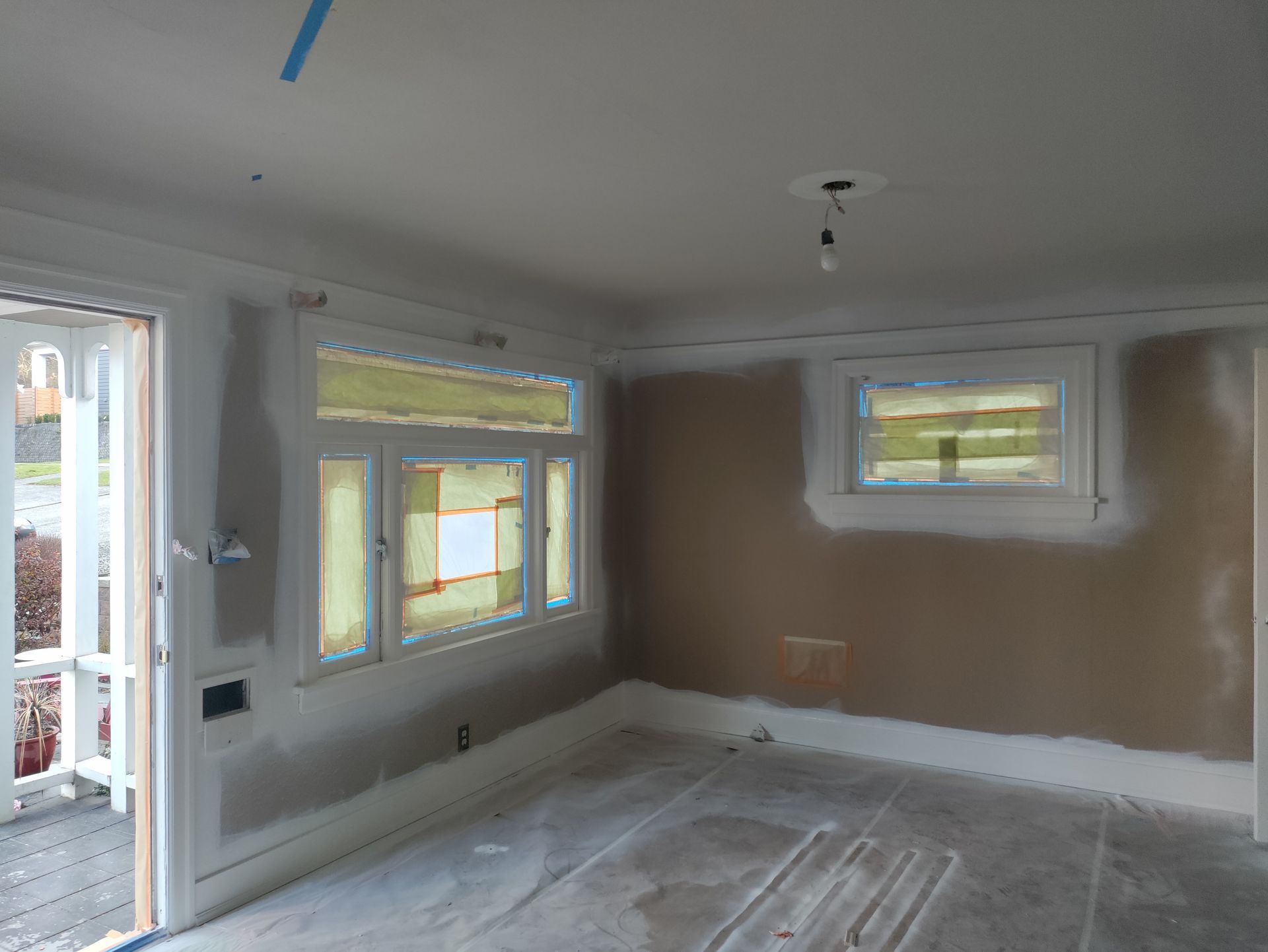 Empty room under renovation with taped windows, white walls, and plastic-covered floor.
