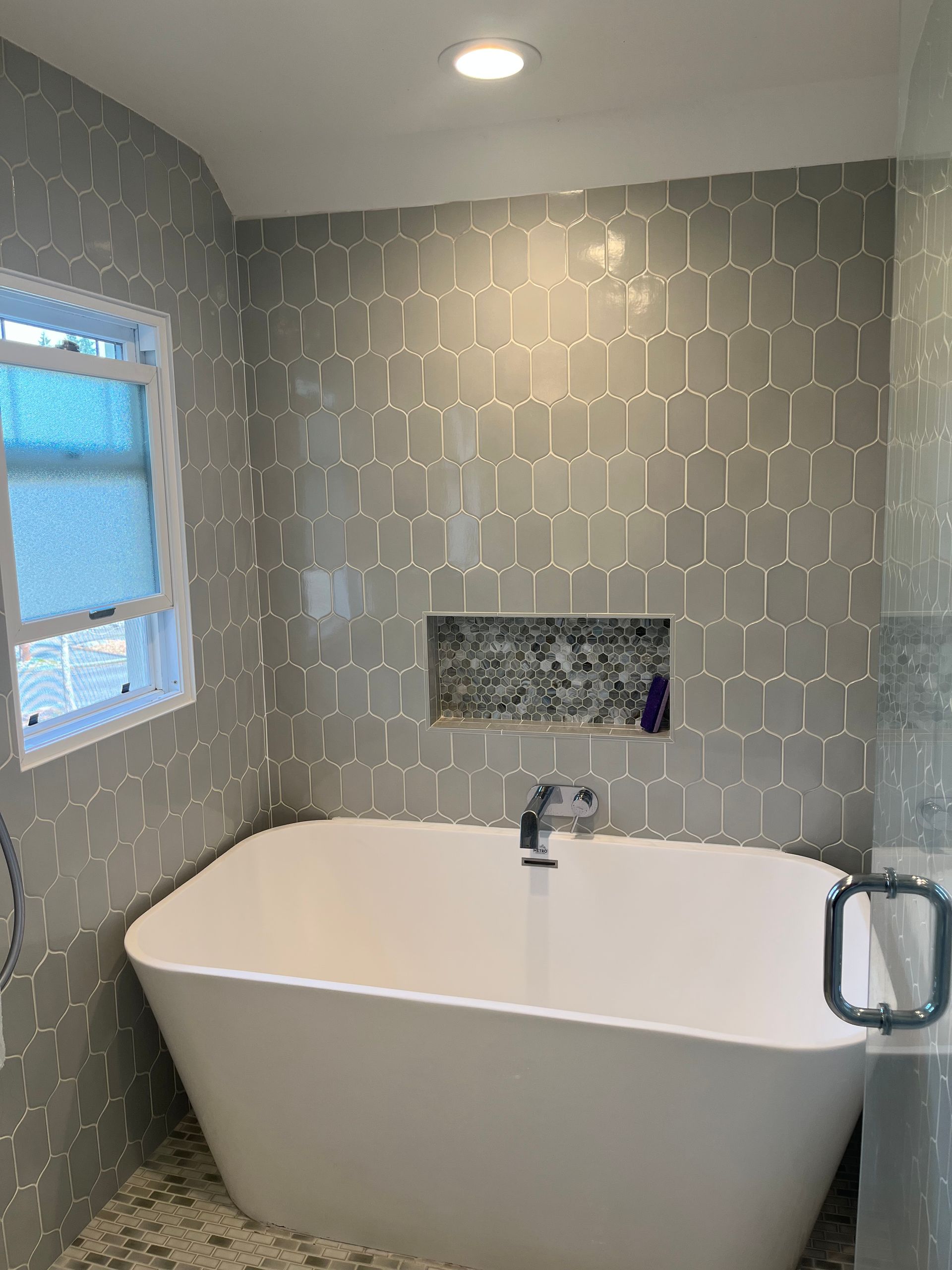 Modern bathroom with freestanding white tub, gray patterned tile, window, and recessed wall niche.