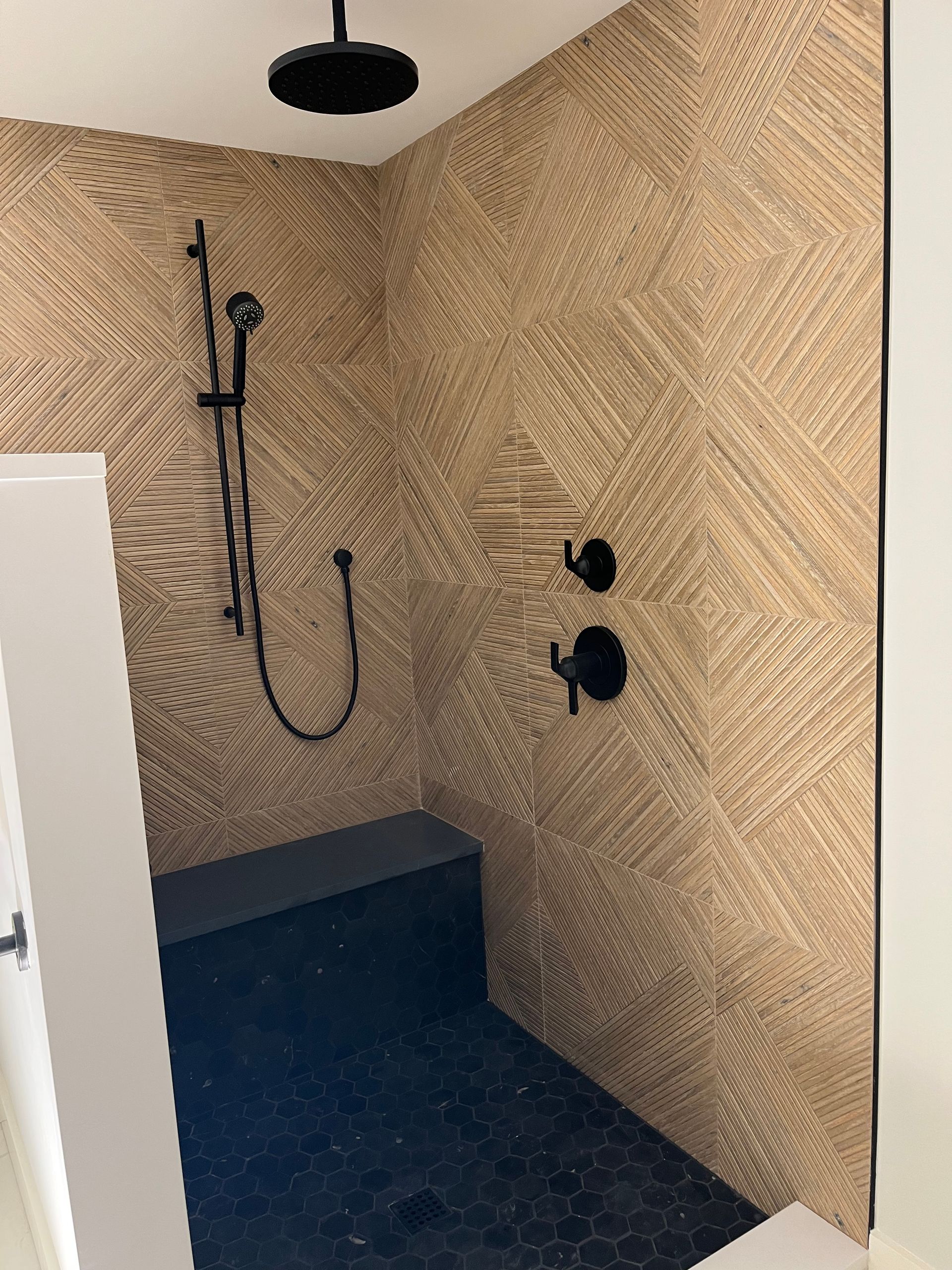 Walk-in shower with woven tan tile, black fixtures, and dark floor in a corner bathroom.