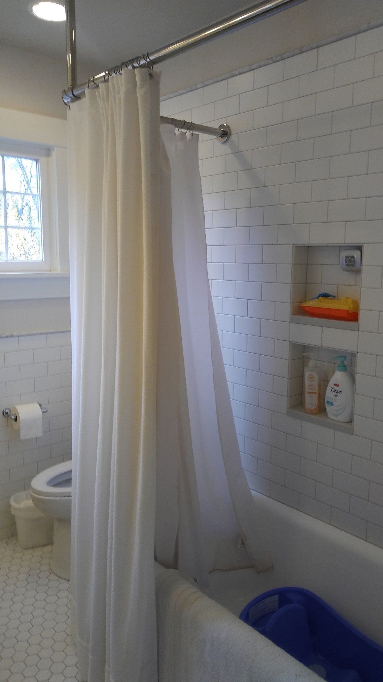 Bathroom with white tile, shower curtain, and bathtub beside a window and wall shelf.