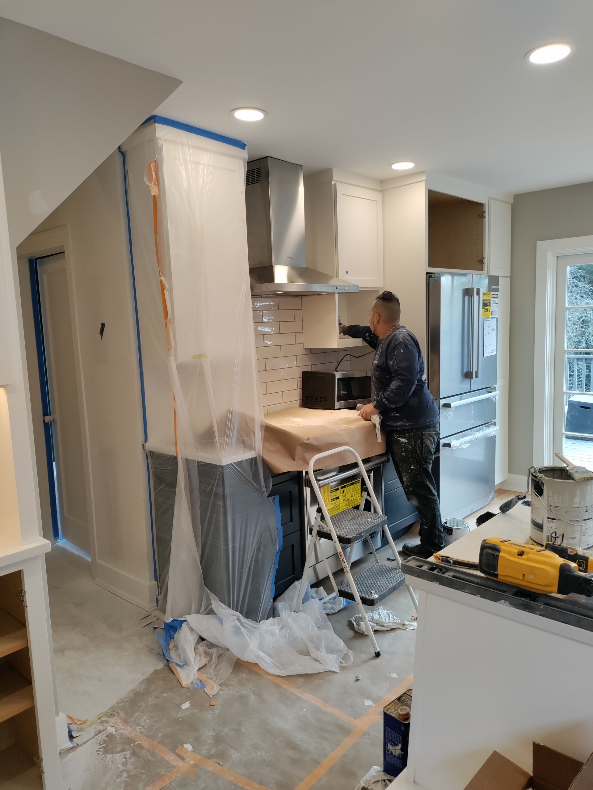 Worker installing kitchen cabinets in a bright home, with plastic sheeting and tools covering the renovation area.