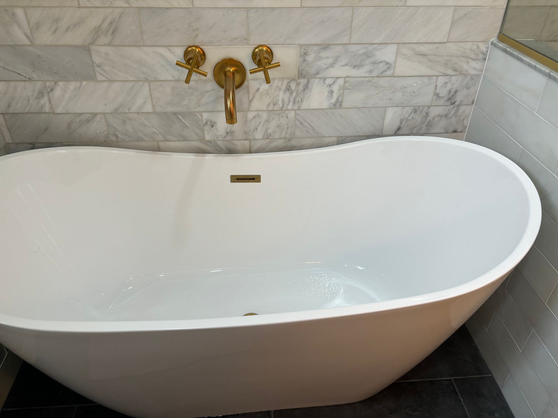 White freestanding bathtub with gold fixtures against a marble-tiled bathroom wall