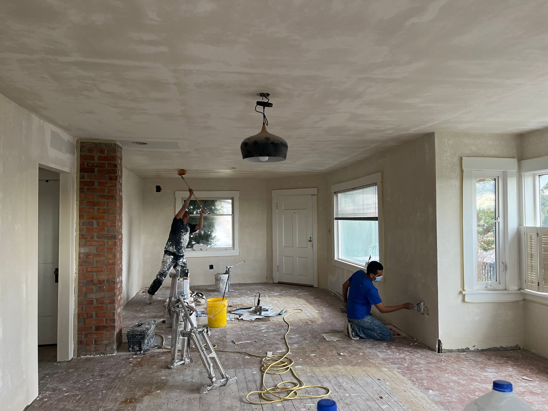 Room under renovation with workers, ladders, tools, and exposed brick wall by bright windows