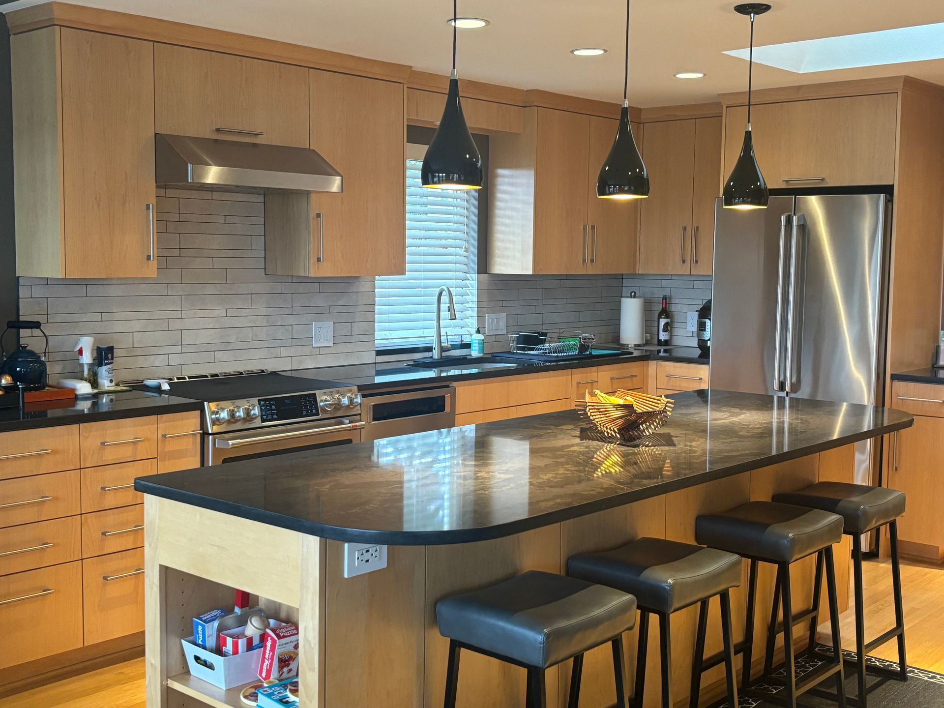 Modern kitchen with island, black stools, wood cabinets, pendant lights, and stainless steel appliances