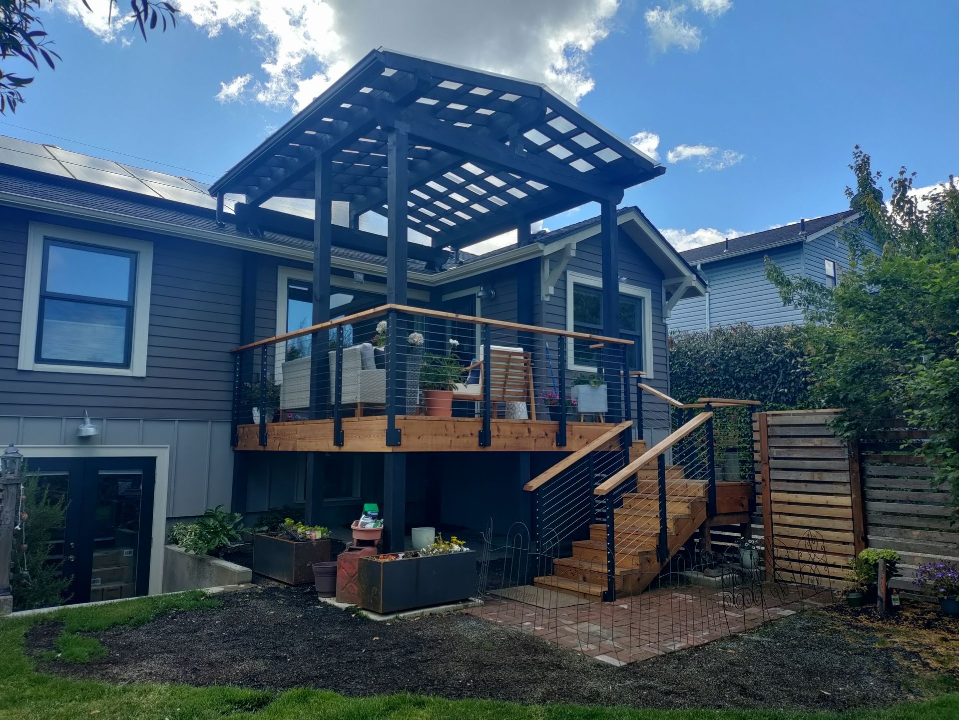 Backyard deck with black railings, stairs, pergola roof, and potted plants beside a house