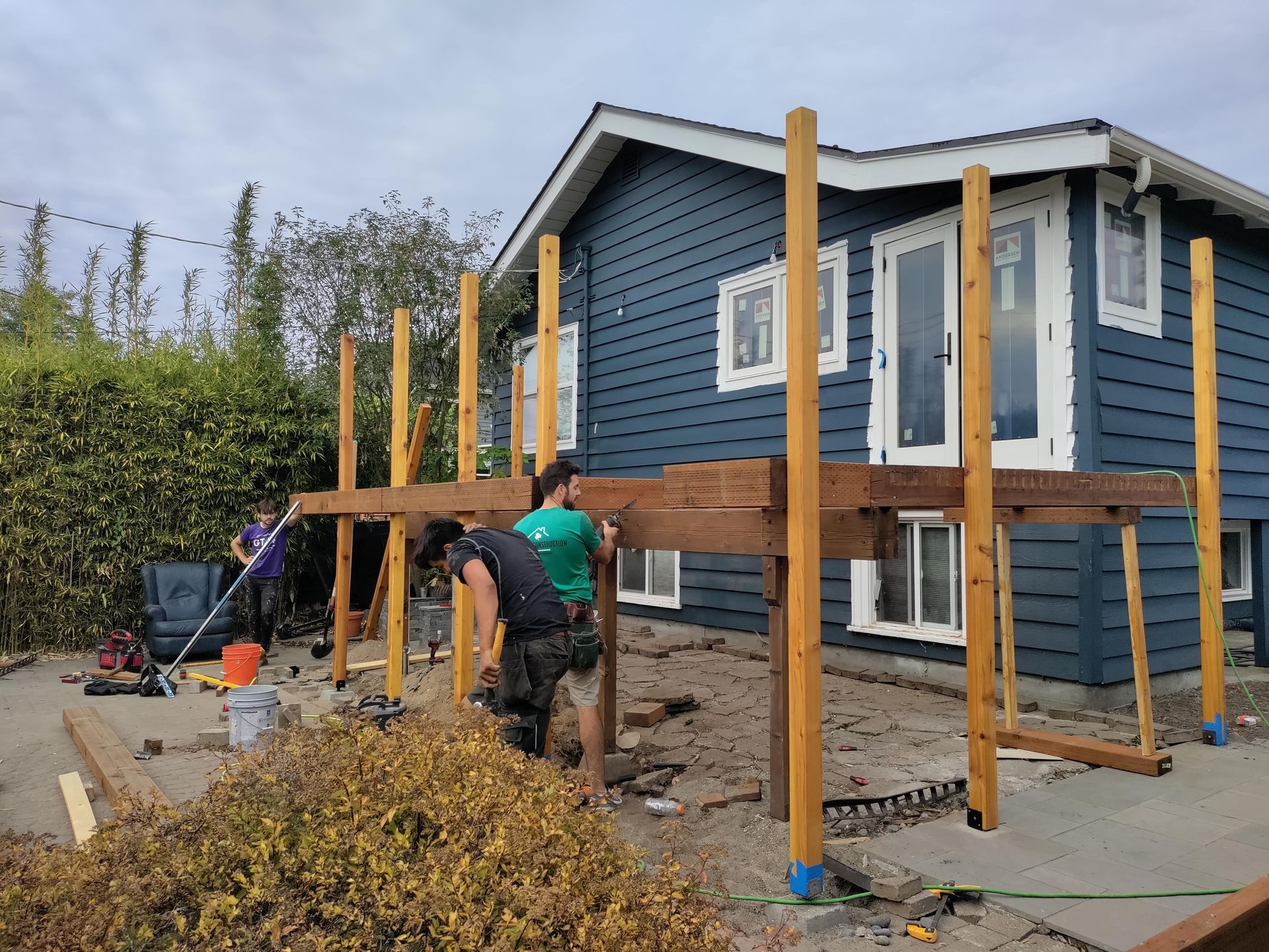 Workers building a wooden deck beside a blue house, with vertical posts and tools on the ground