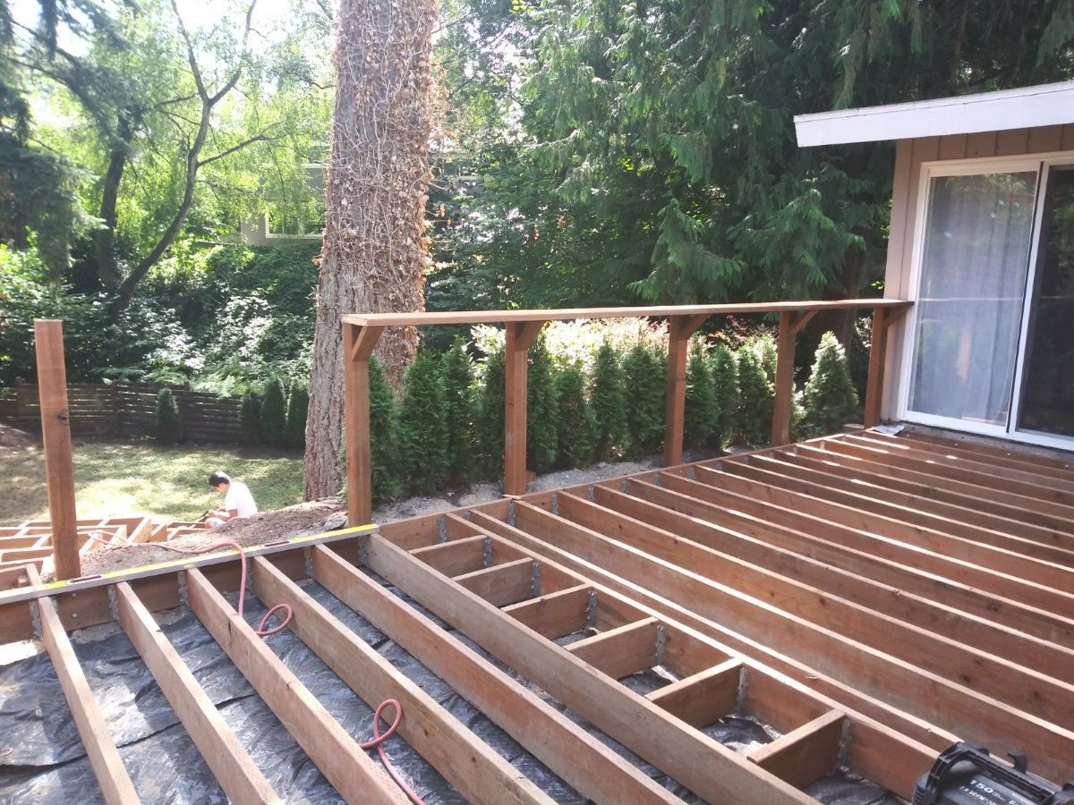 Wooden deck under construction beside a house, with exposed joists and railing posts in a wooded yard