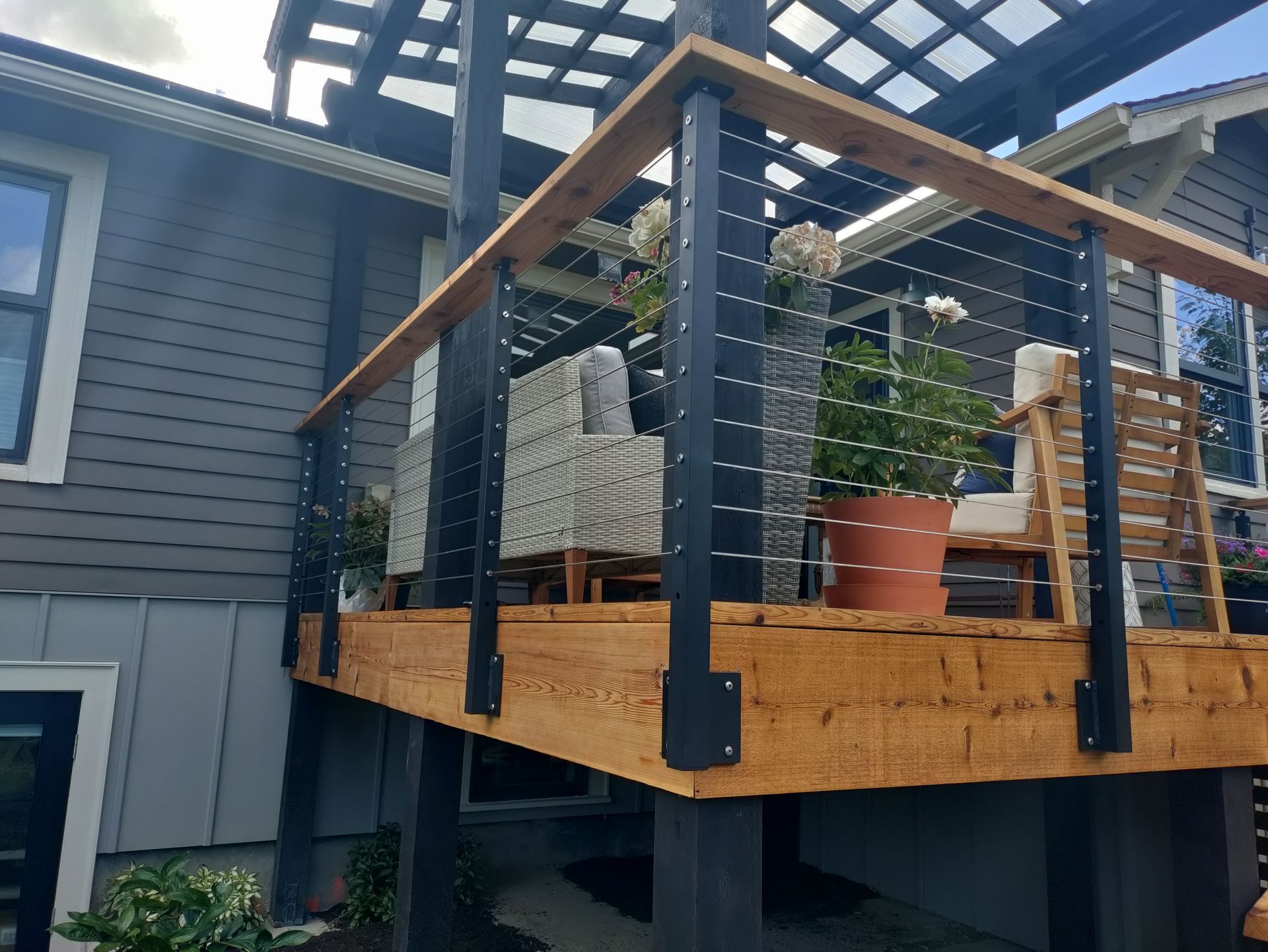 Small raised deck with railing and potted plants beside a gray house under a pergola