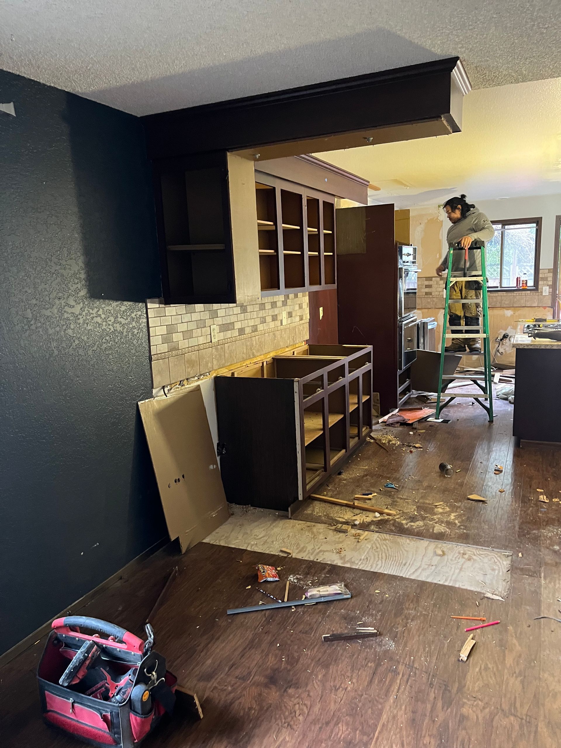 Damaged kitchen with broken cabinets, scattered debris, and a person working near the window.