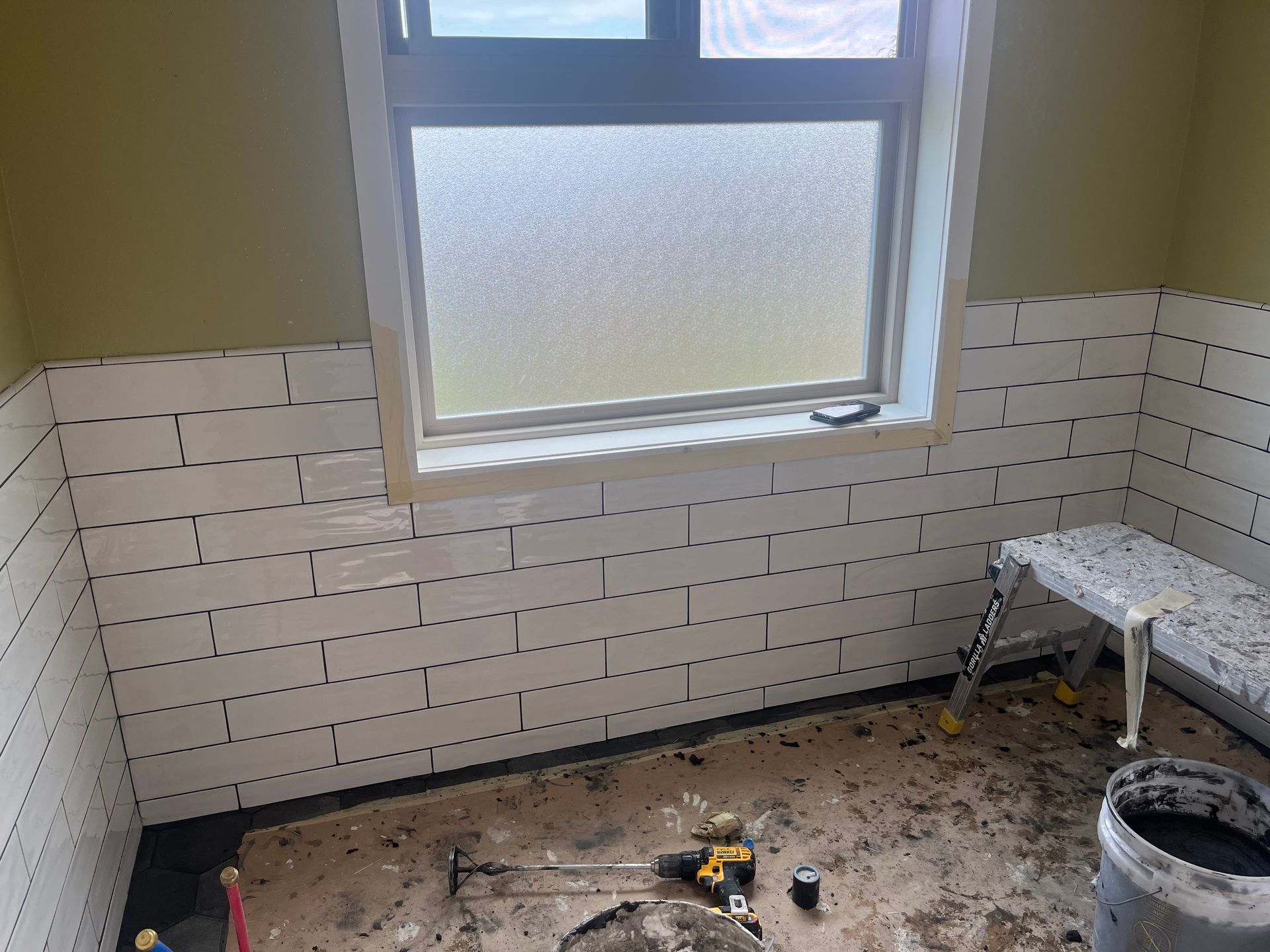 Partially tiled bathroom under renovation with a window, tools, debris, and a bucket on the floor