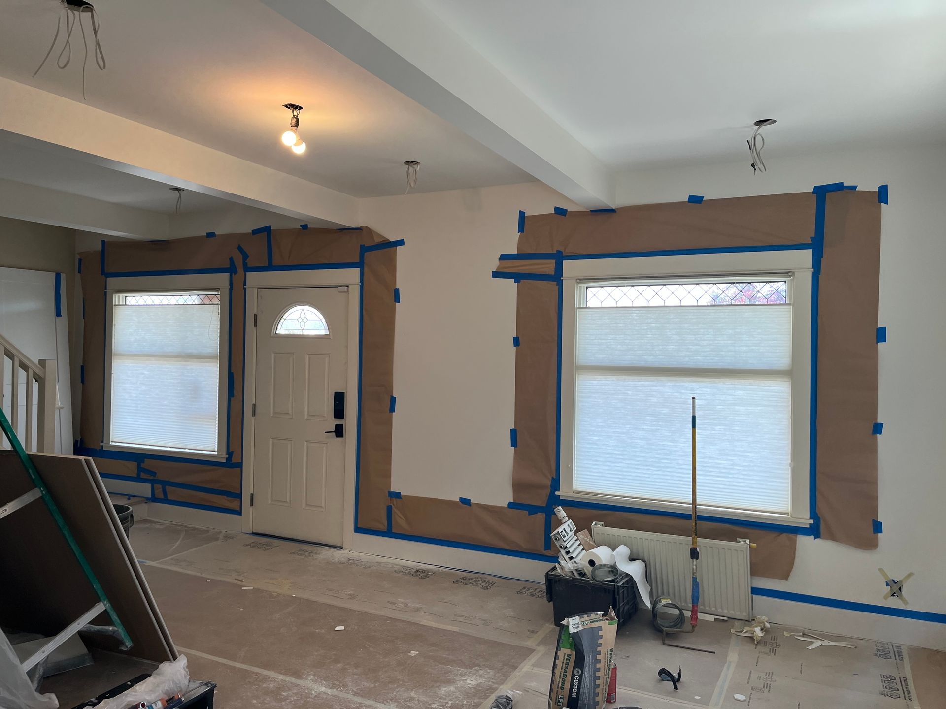 Room under renovation with taped windows and door, exposed trim, and construction tools on the floor.