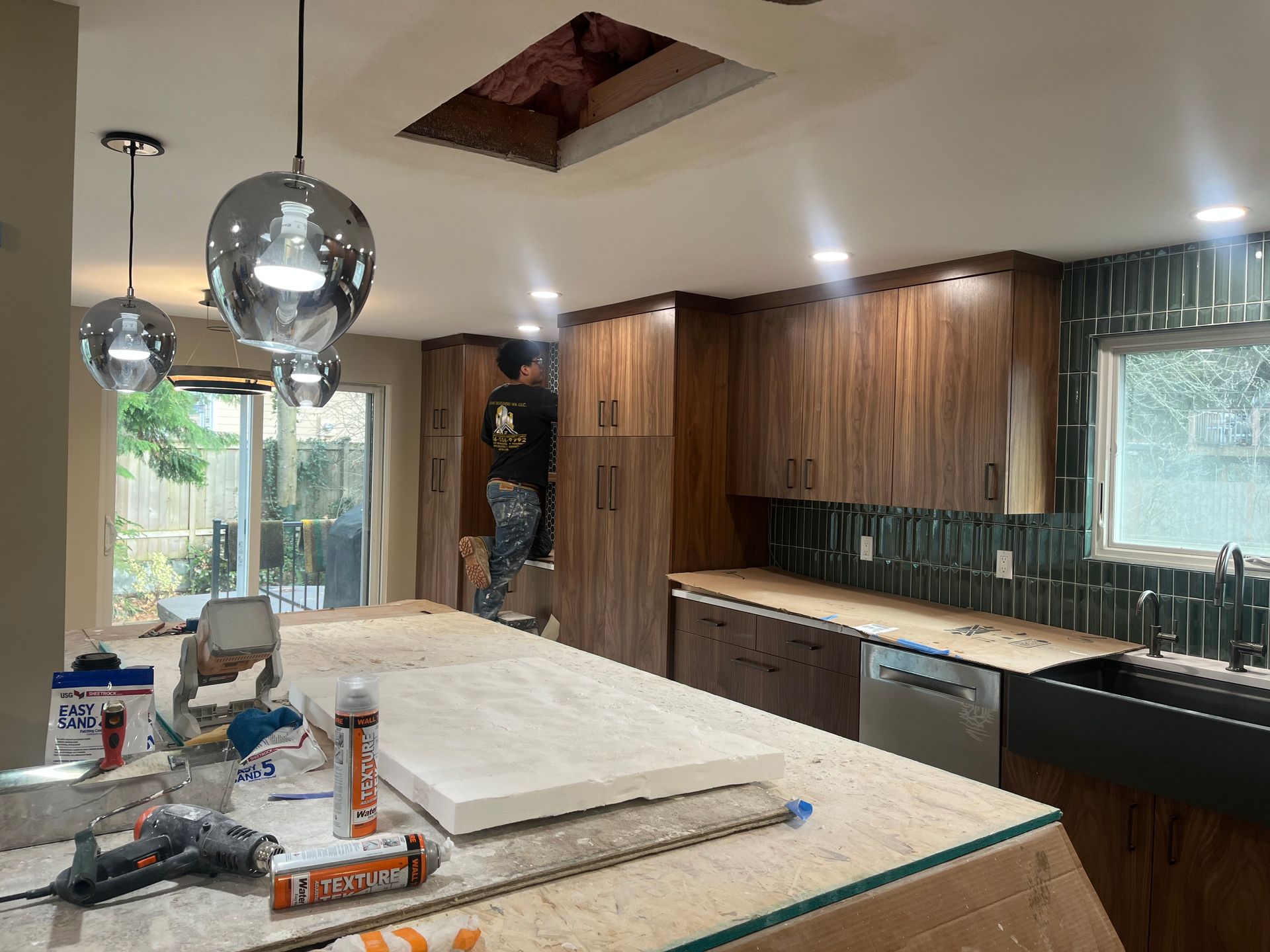 Kitchen under renovation with wood cabinets, countertop tools, and a worker near the back wall
