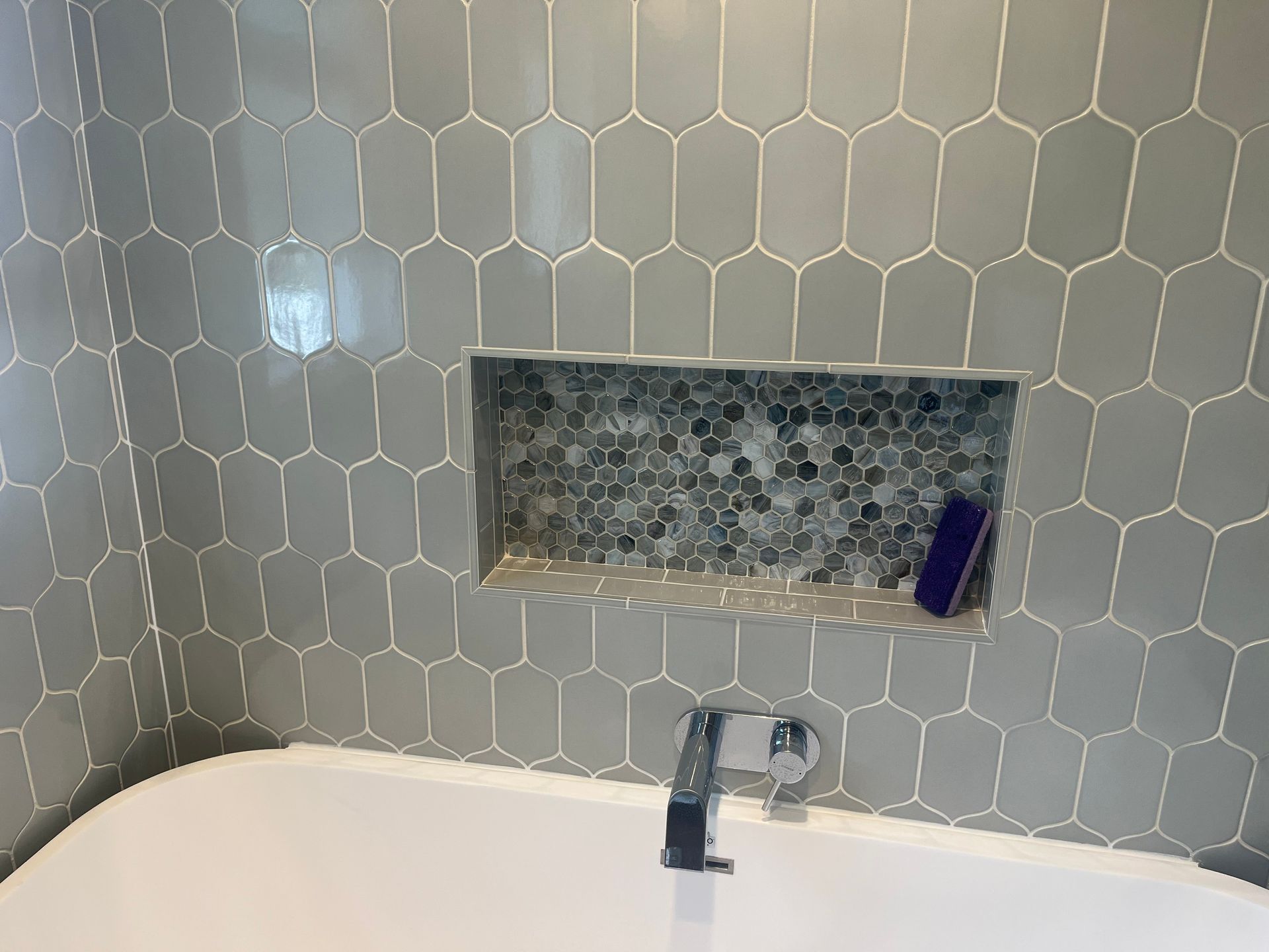 Bathroom tub with gray hex tile wall, recessed shelf, and chrome faucet.
