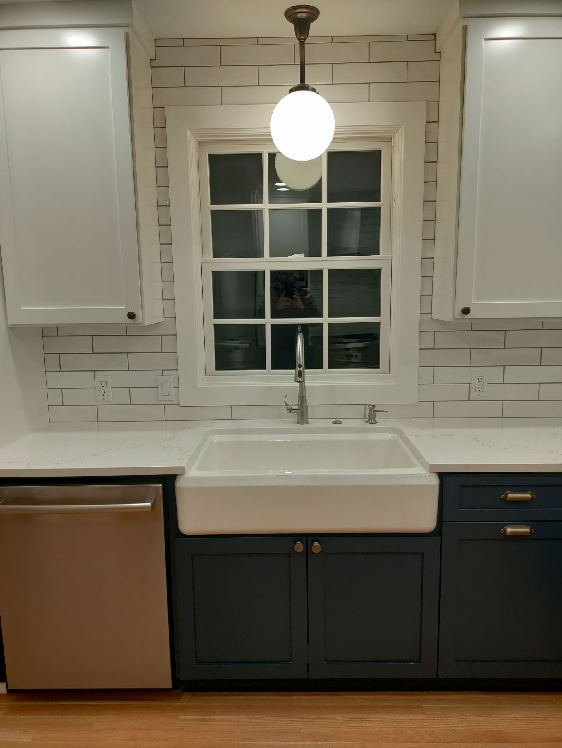 Modern kitchen sink beneath a window with white tile backsplash, brass pendant light, and navy lower cabinets