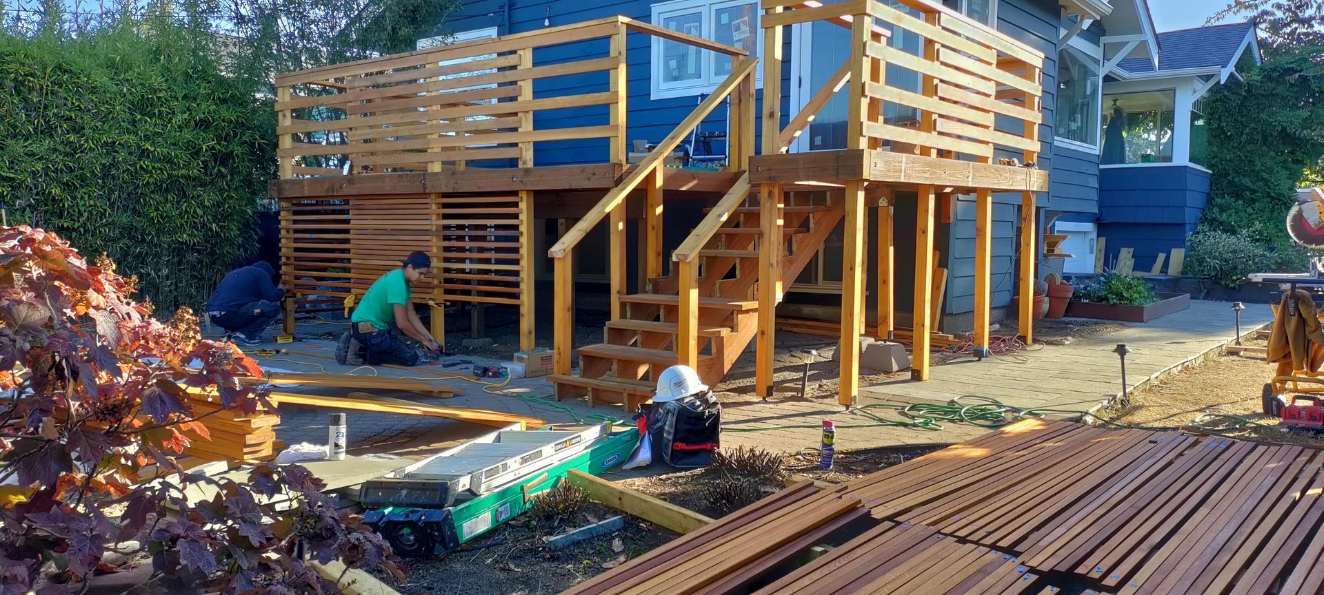 Workers building a wooden house frame at a sunny construction site, with lumber stacked nearby.