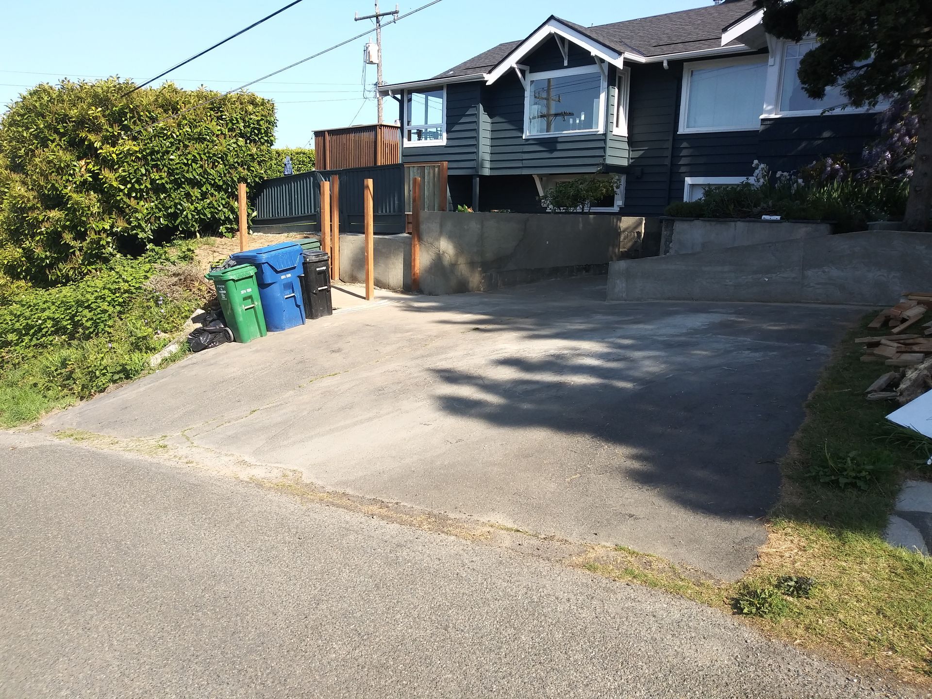 Driveway beside a house with two trash bins, a wooden gate, and bright sunlight casting shadows.