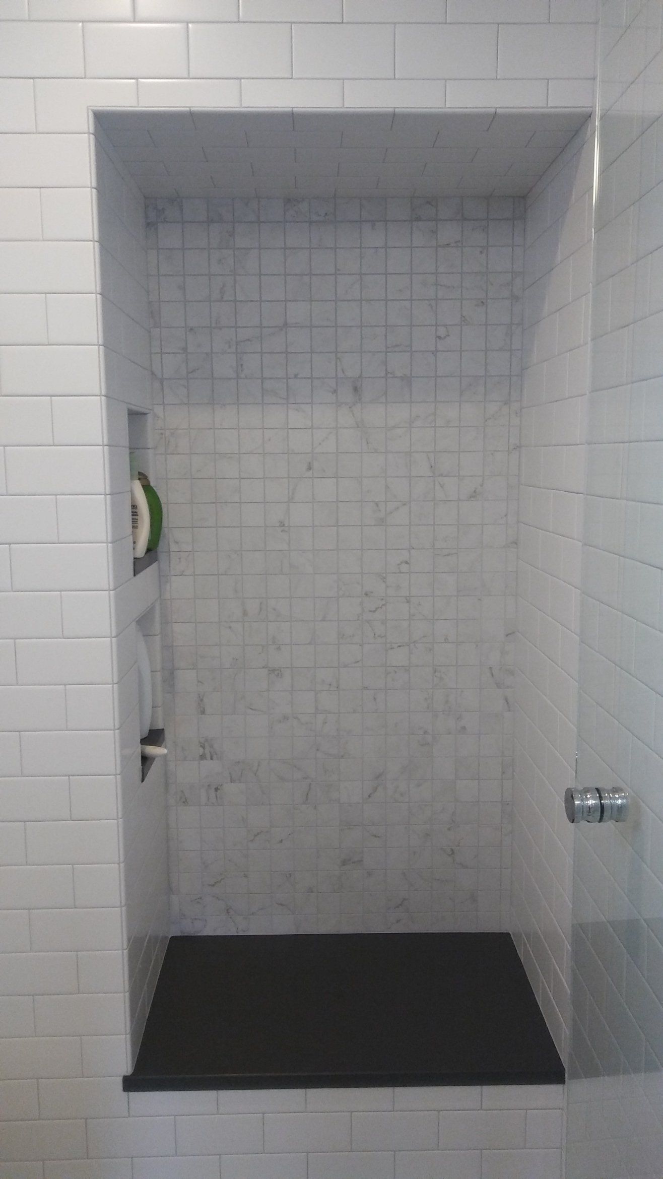 Small tiled shower stall with a dark floor mat and open door.