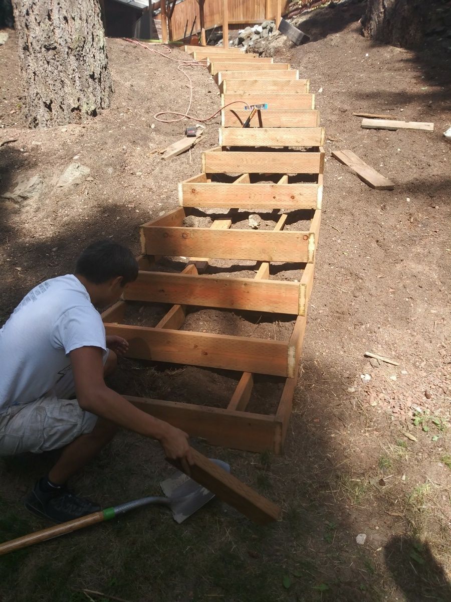 Person building a wooden staircase outdoors on a dirt slope using tools and planks